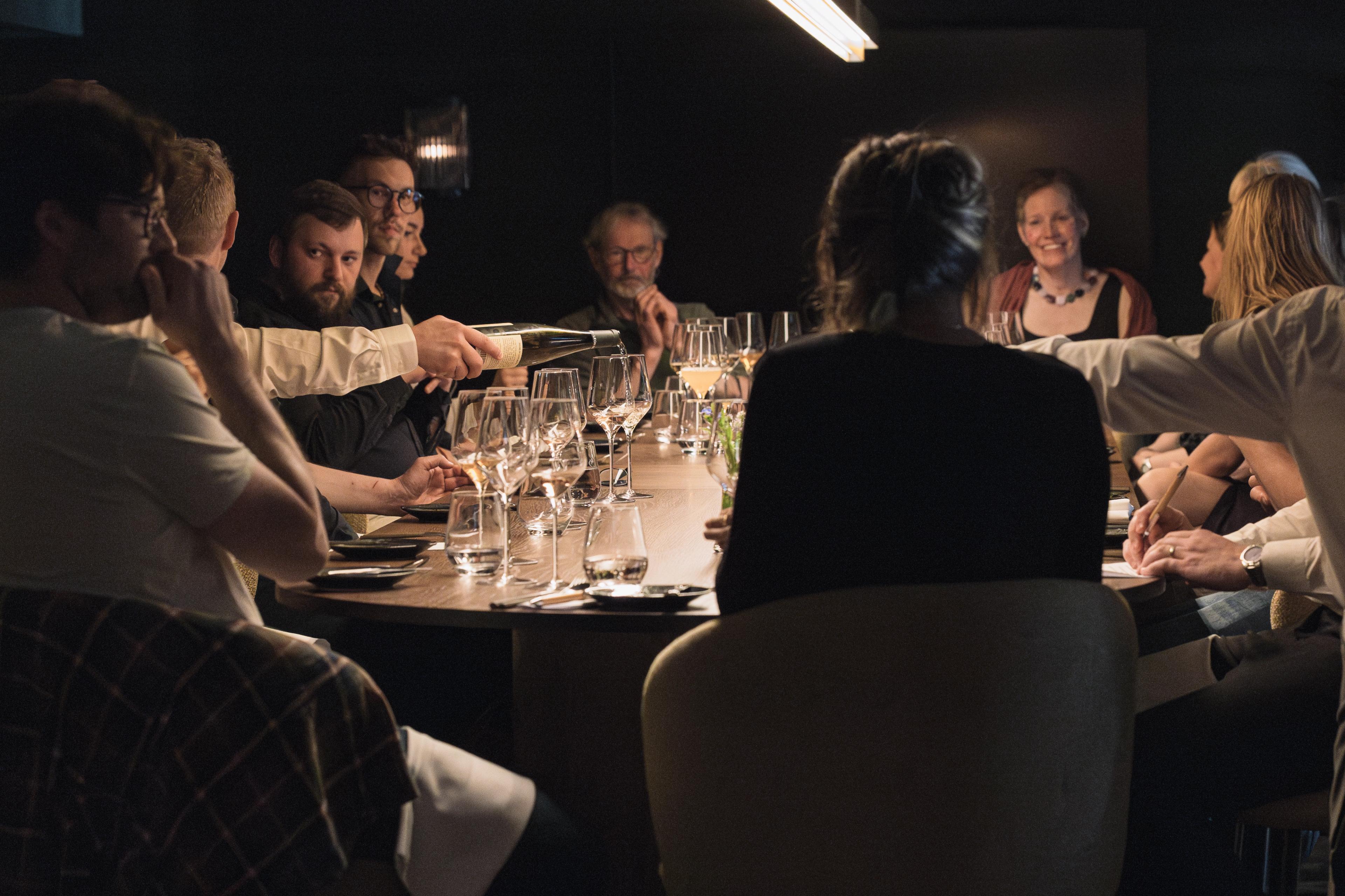 Group of people sitting aroud a big dining table. Wine in beeing poured into the wine glasses.