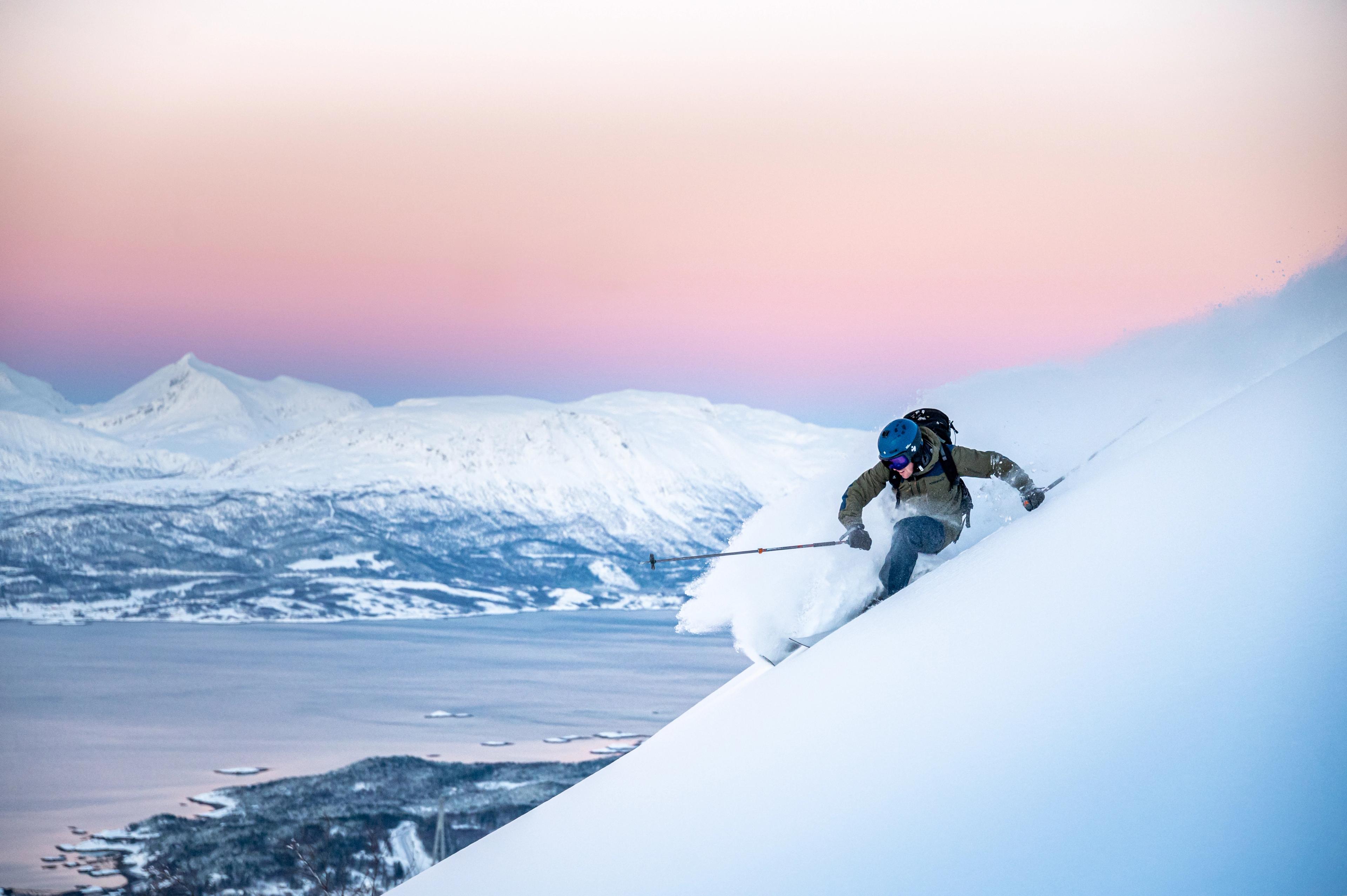 Skier making fresh tracks in deep powder above the fjord in Narvik. Pink sky behind snow covered mountains. 