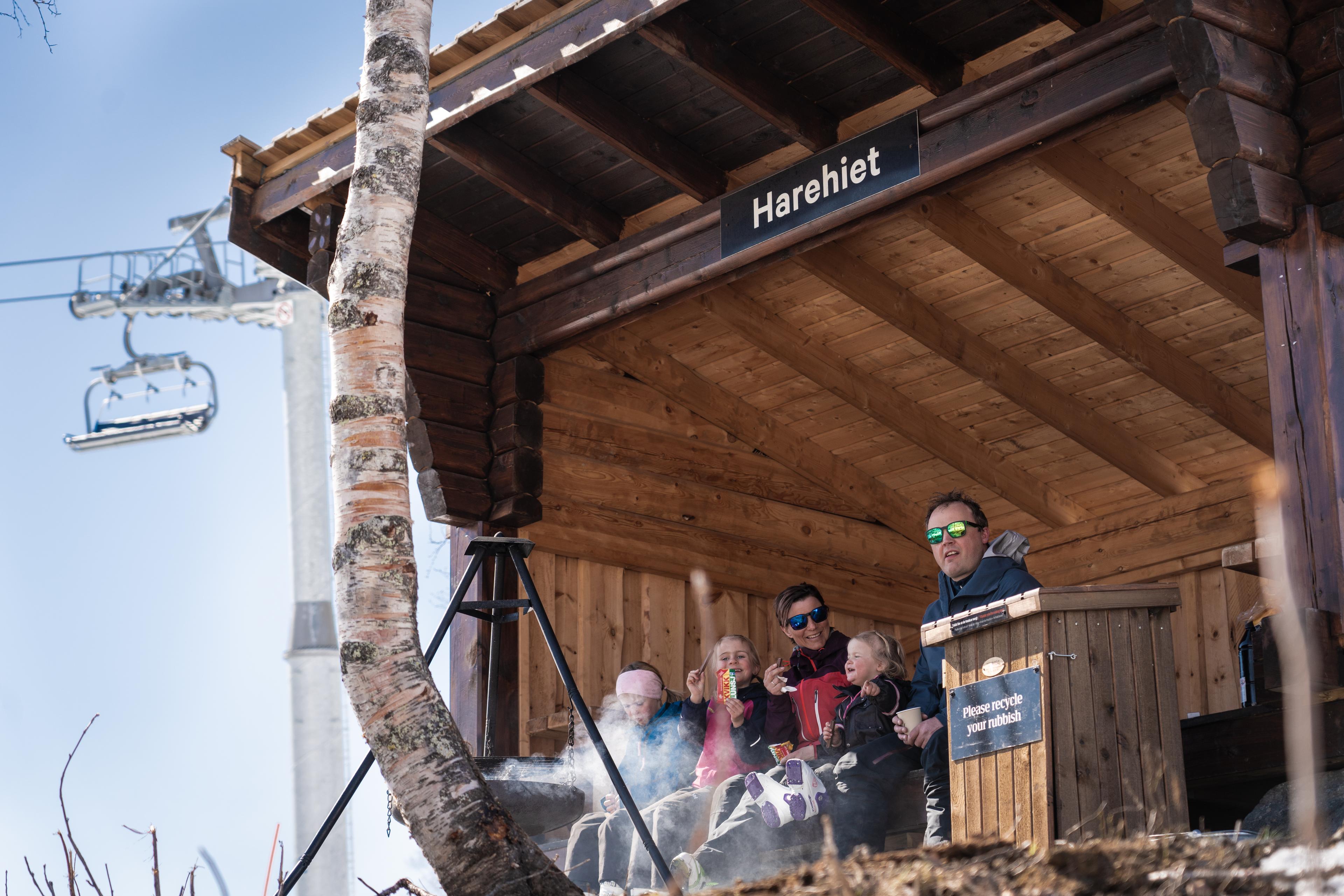 Familie koser seg med grilling i gapahuken Harehiet i solskinn, med stolheisen i bakgrunnen.