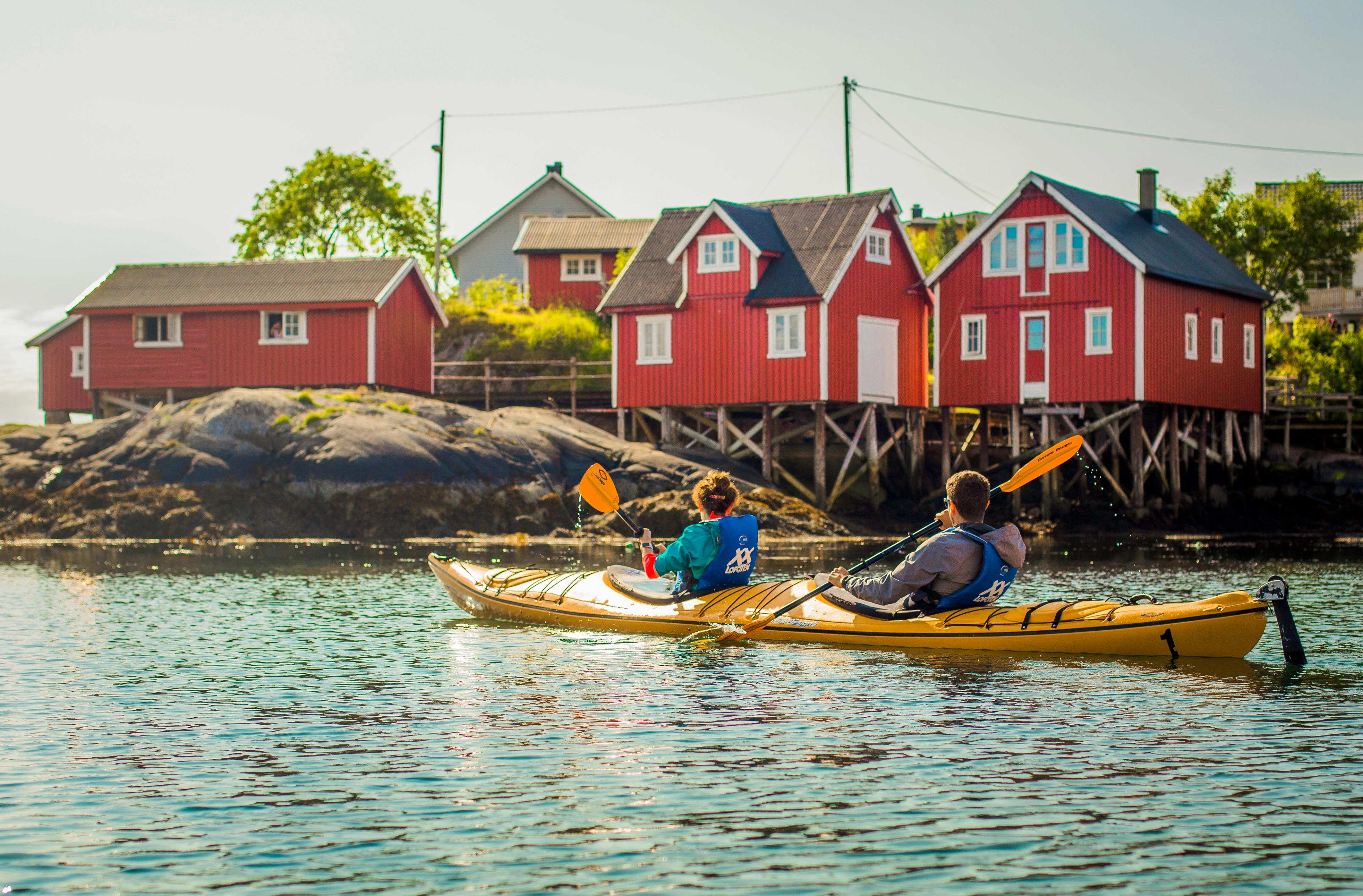 Man and woman paddling together in a kayak and looking up at some red fishermen's huts
