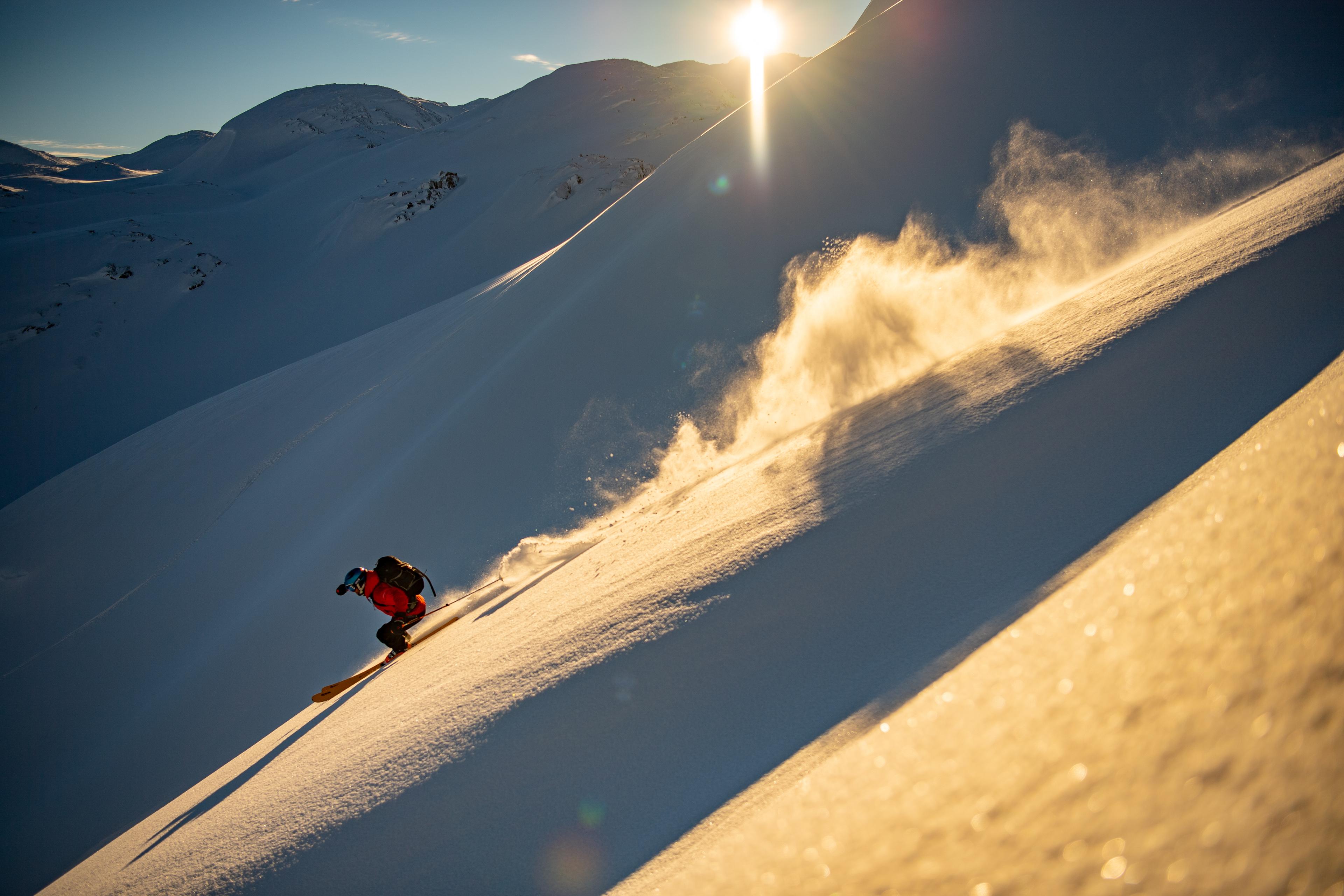 Frikjører står ned bratt bakke så snøføyka står bak han. Sola titter så vidt over fjellene i bakgrunnen.