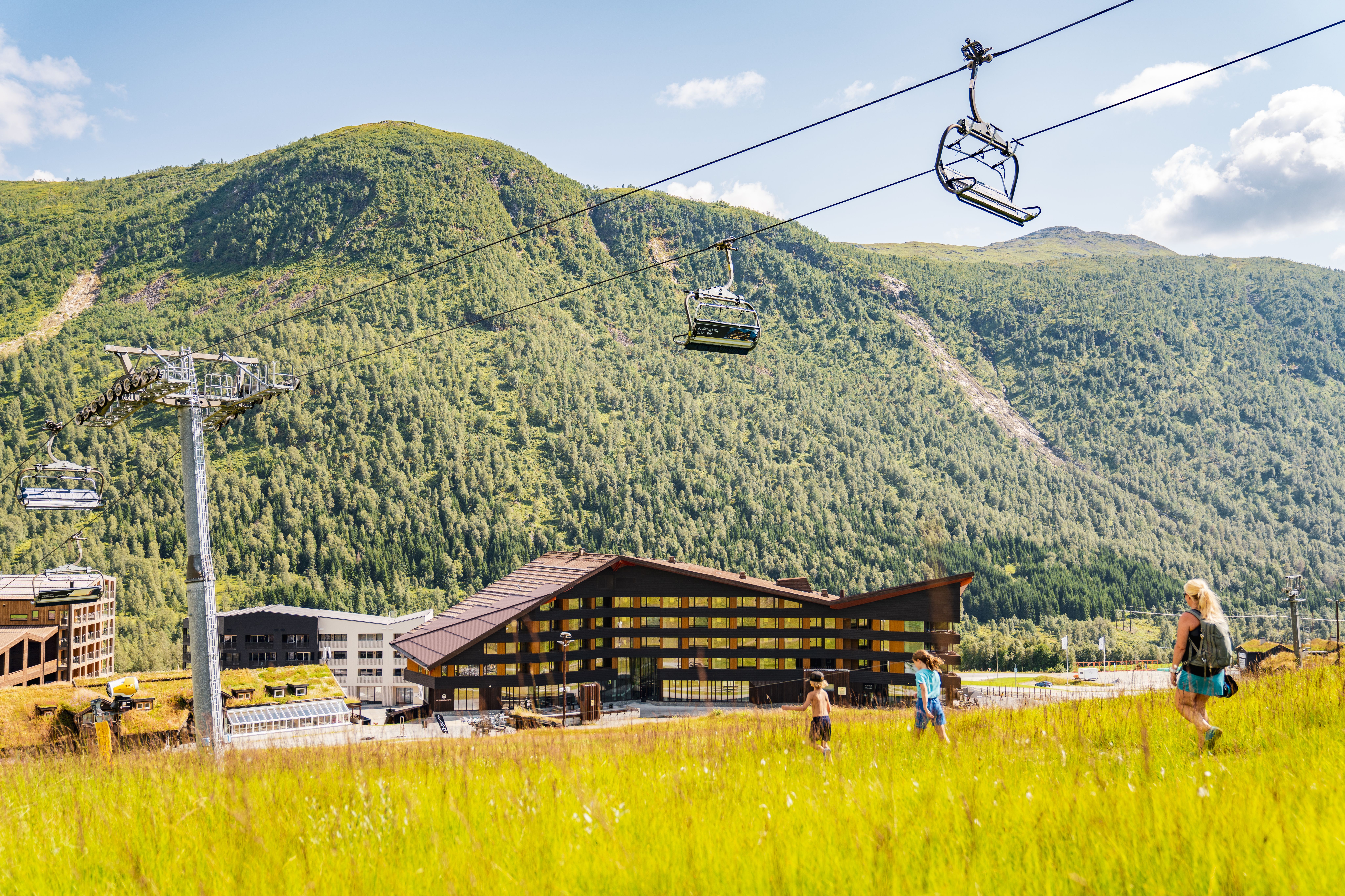 A woman and two children walk in the green grass under the chairlift in Myrkdalen on a nice summer day. Myrkdalen Hotel and apartment building seen in front of Svolefjellet.