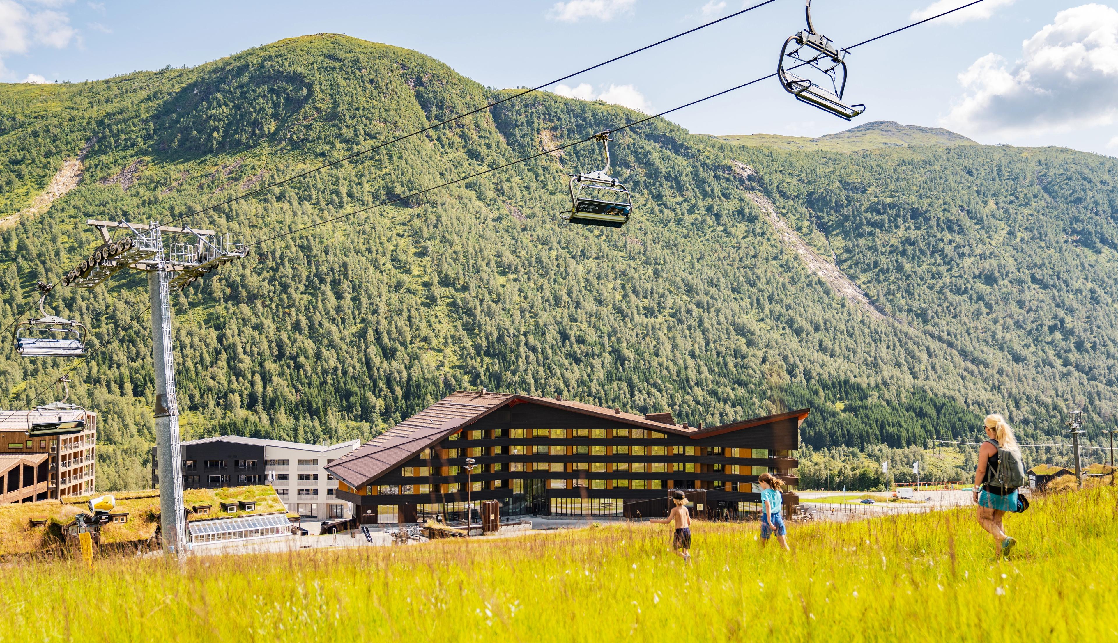 En kvinne og to barn går i grønt gress under stolheisen i Myrkdalen en fin sommerdag. Myrkdalen Hotel og leilighetsbygg sett foran Svolefjellet.