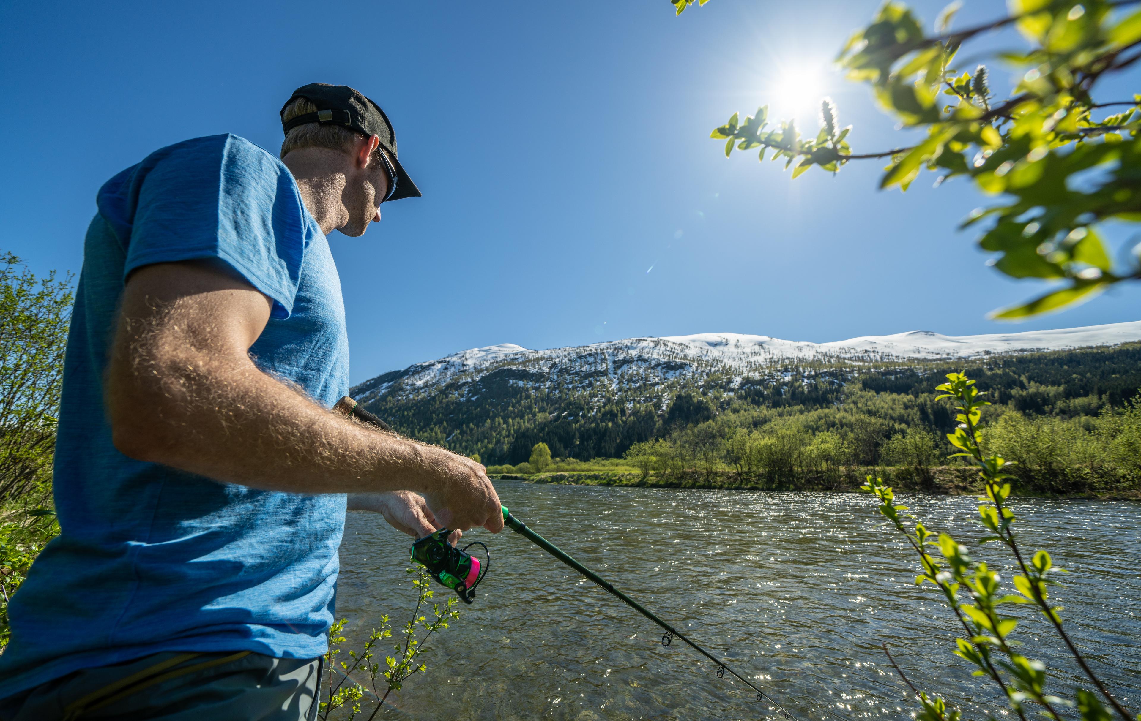 Mann fisker med fiskestang i et fjellvann under blå himmel og solskinn.
