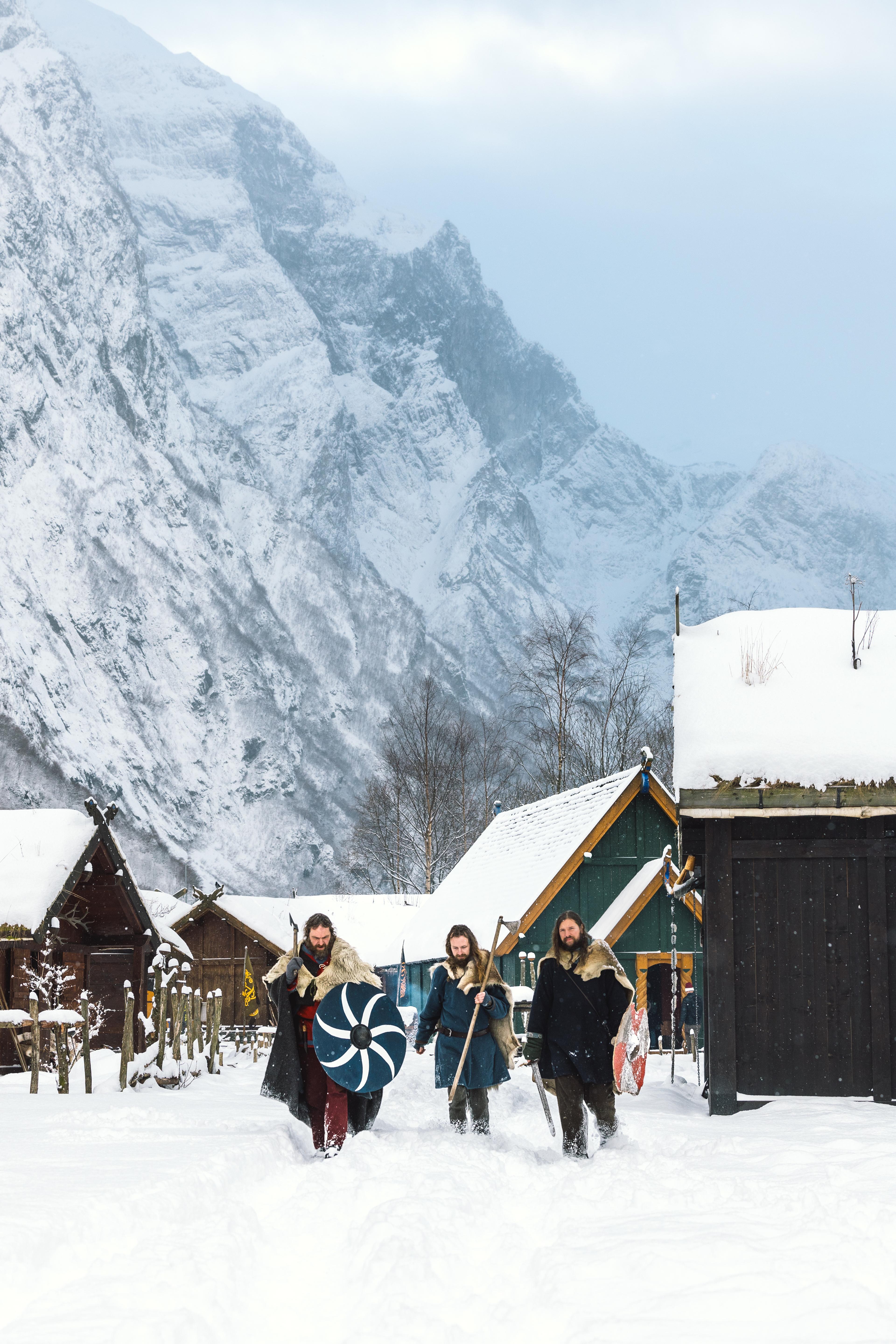 Tre vikingar med skjold og øks går gjennom vikinglandsbyen i snø.