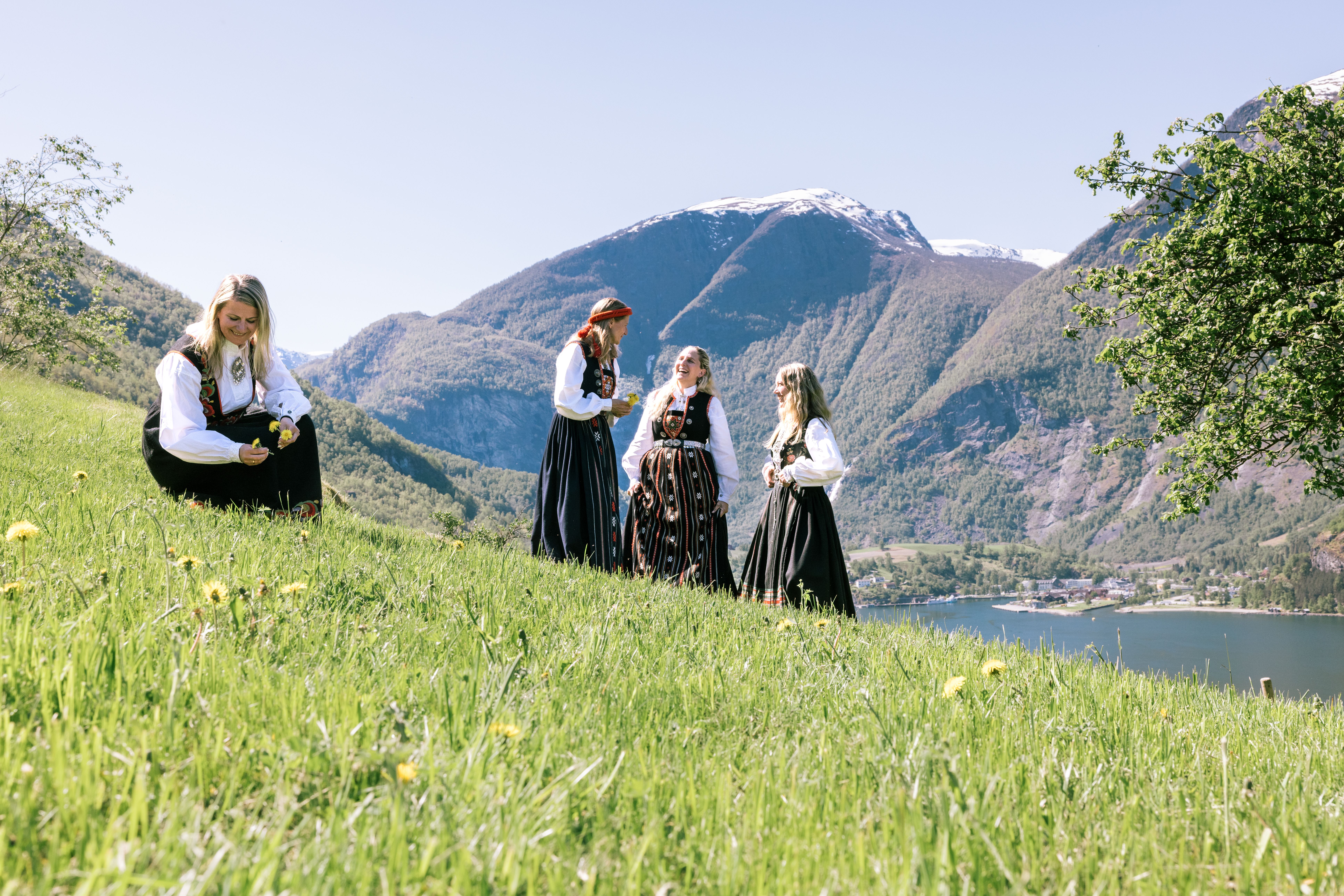 Four women wearing traditional Norwegian bunad stand and sit on a sunny grassy hillside, picking flowers and chatting, with a fjord and steep mountains in the background