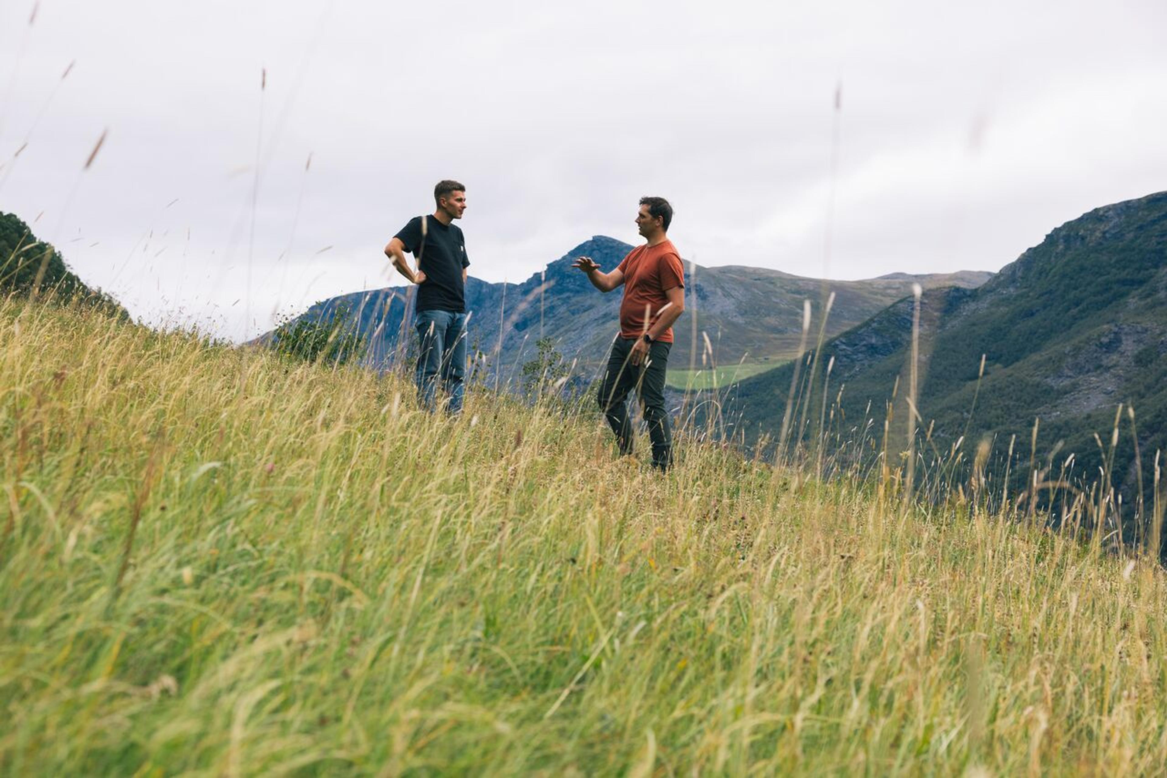 Two men talking in a field, with mountains in the background.