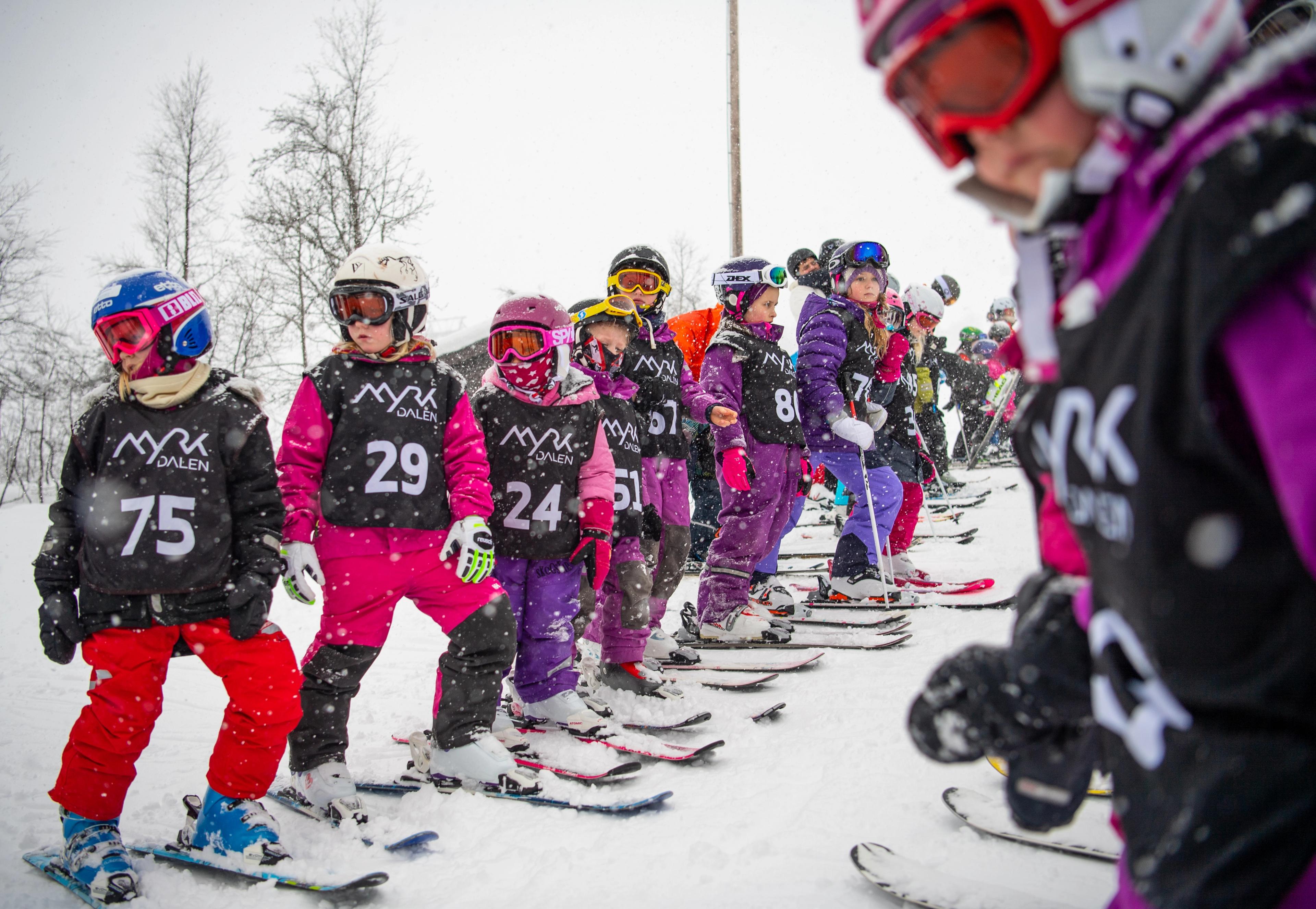 Children in ski gear with race bibs on their chests stand in a row, ready for the ski race.