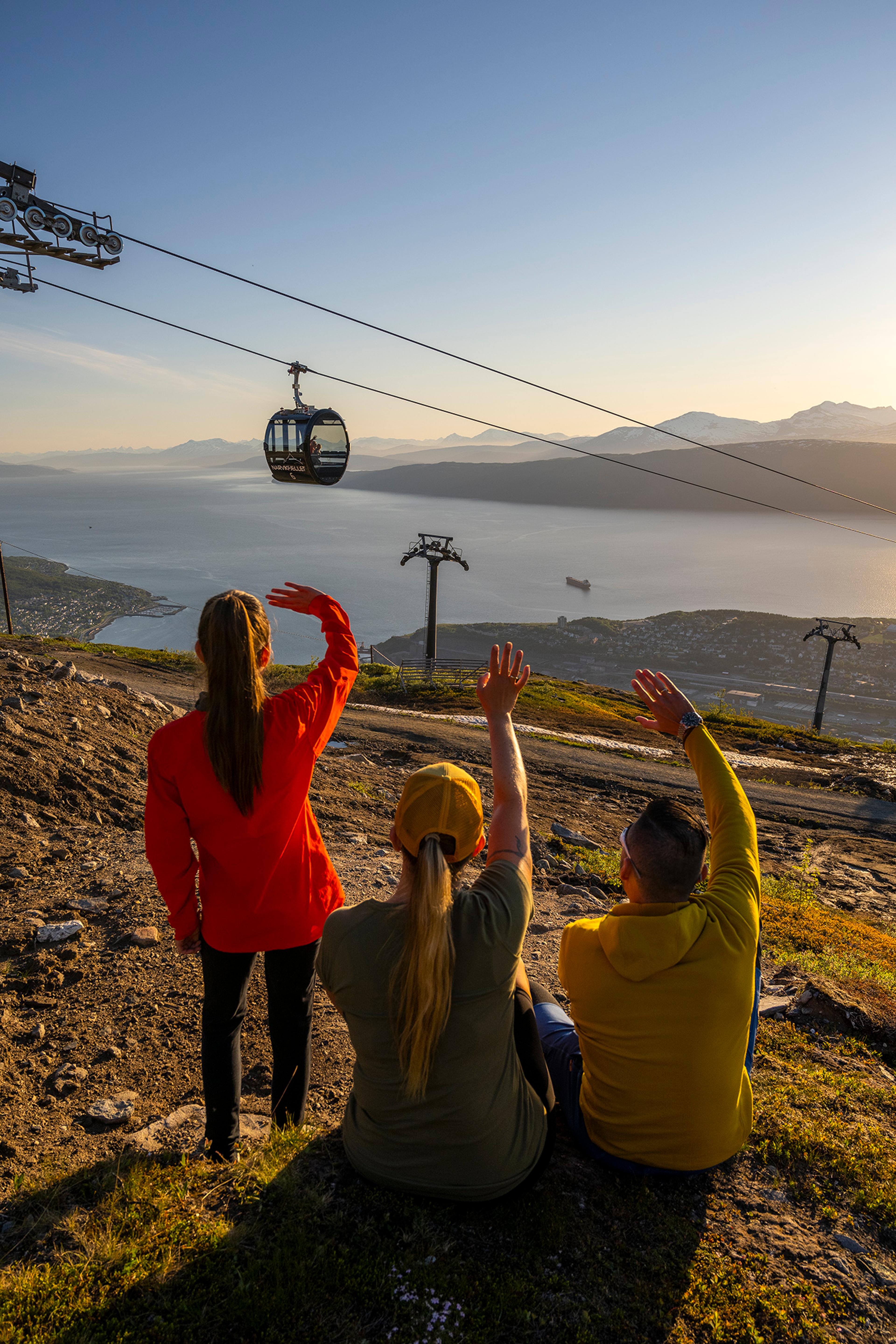 Three people sitting and standing on a mountainside wave towards the Narvik Cable Car, with the Ofotfjord in the background.