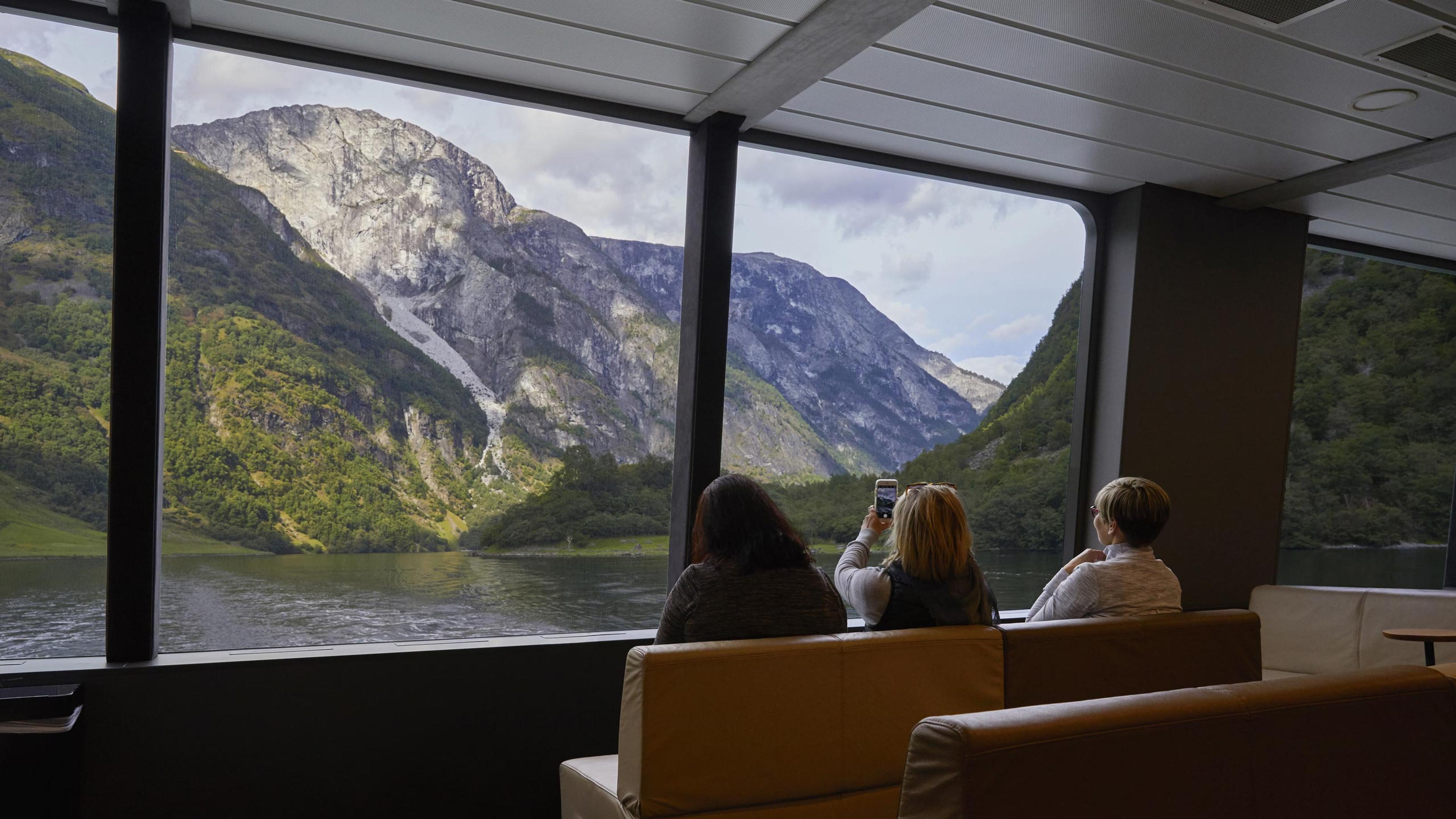 Three people seated indoors on a boat during a fjord cruise in Nærøyfjord in Norway, looking out through large windows at a stunning landscape with steep, rocky mountains and lush greenery. One person is capturing the view with a smartphone.