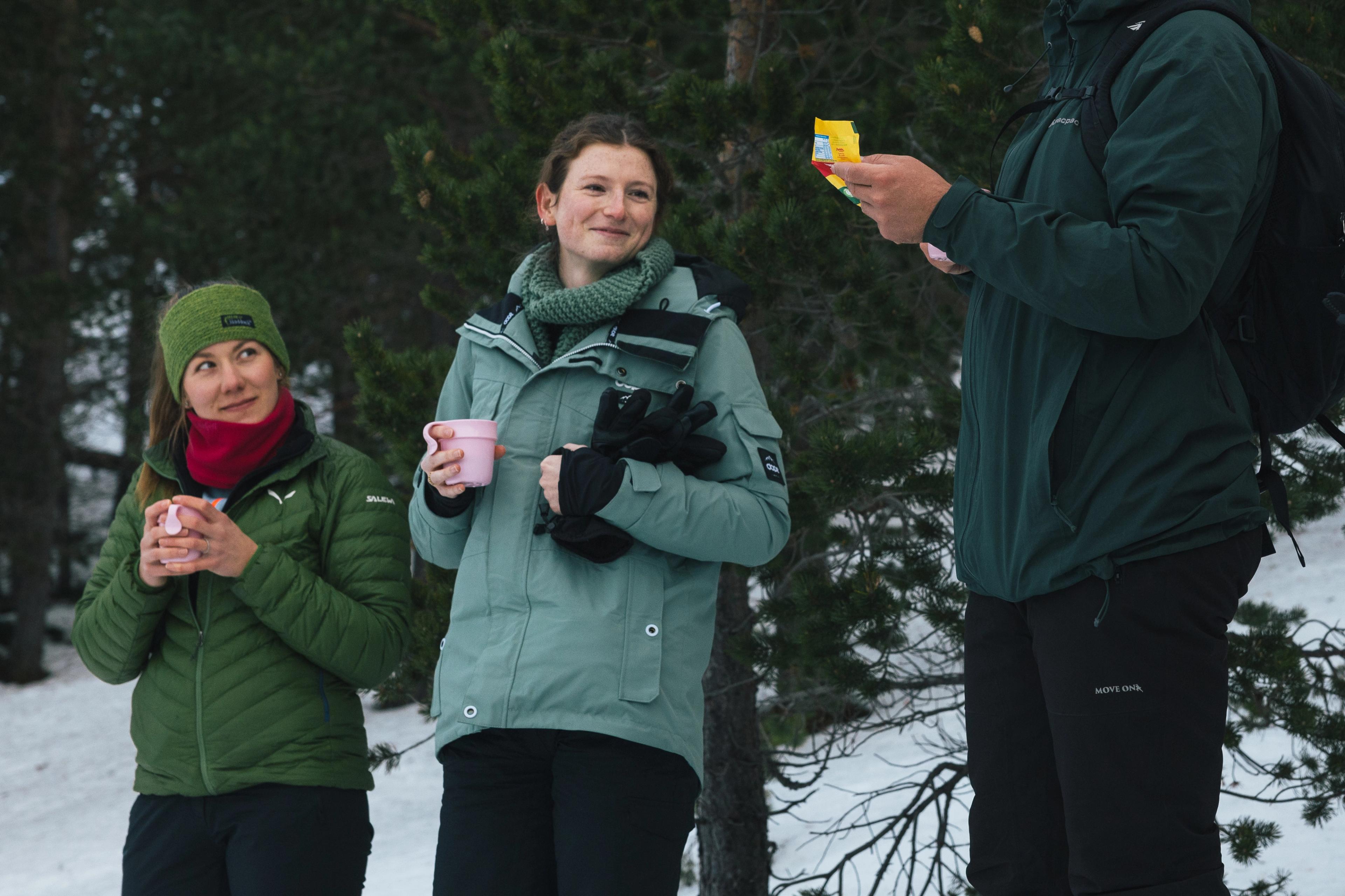  People in winter clothing appear to be enjoying hot drinks and quick lunches in the forest.