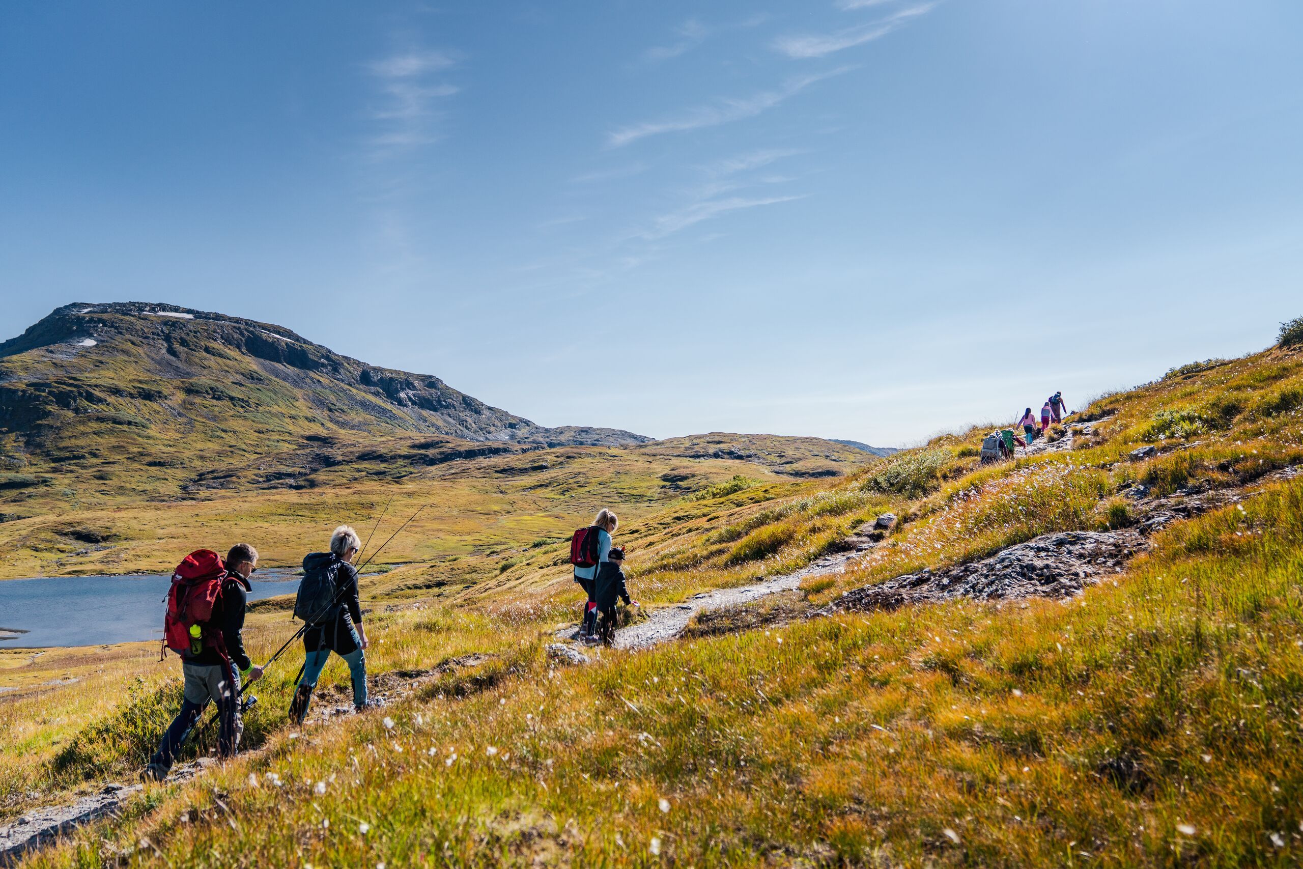 En kvinne og to barn går i grønt gress under stolheisen i Myrkdalen en fin sommerdag. Myrkdalen Hotel og leilighetsbygg sett foran Svolefjellet.