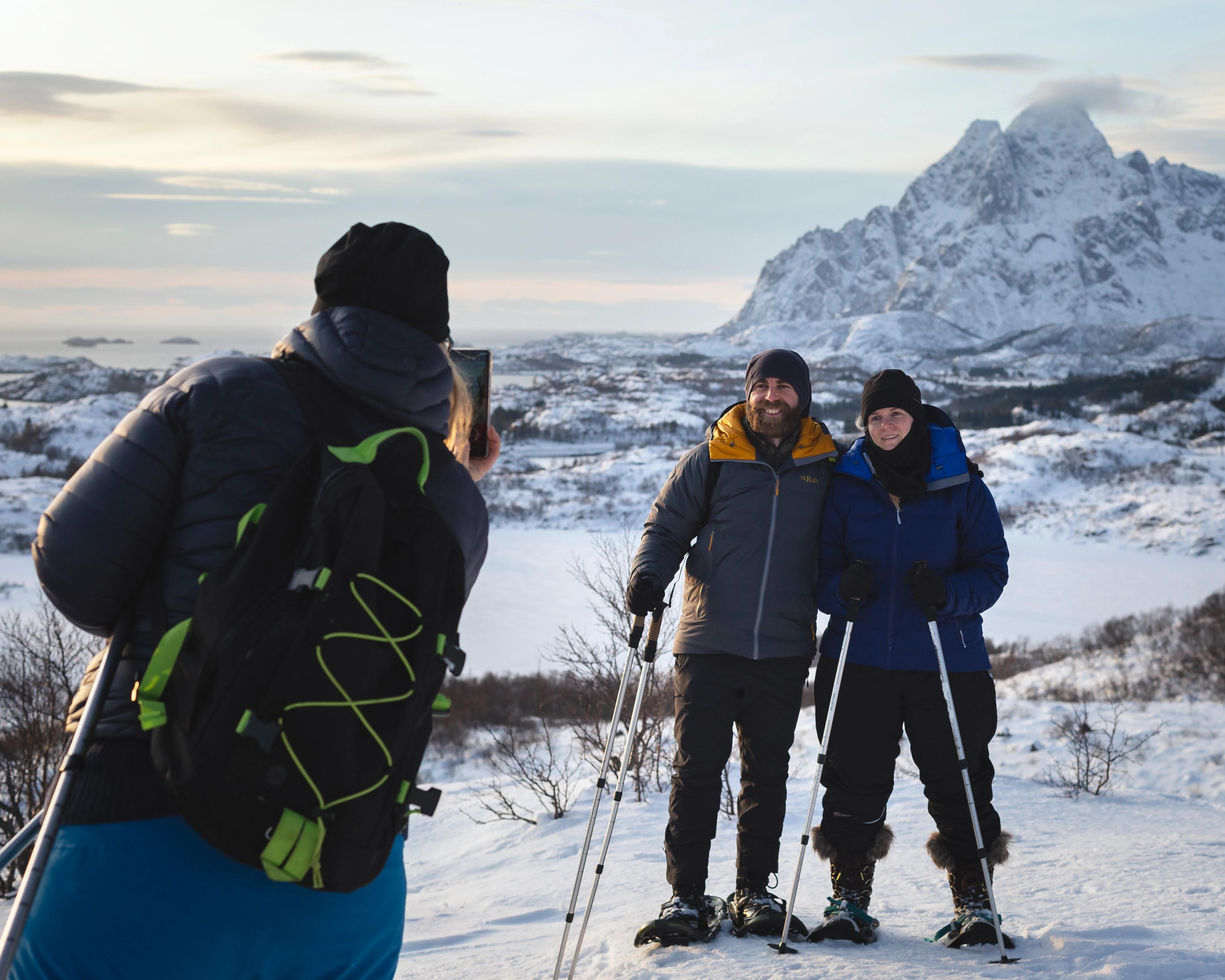 A guide captures a couple on snowshoes and poles standing before a mountain summit.