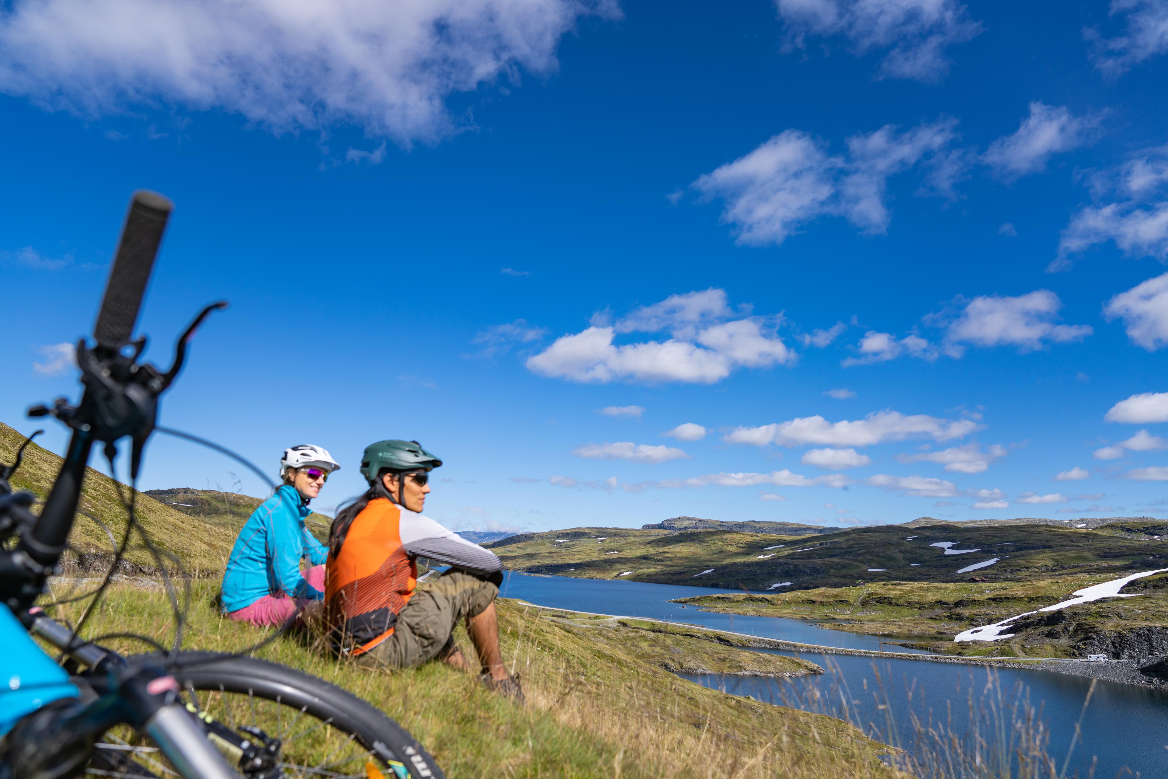 To kvinnelige syklister tar seg en pust i bakken og sitter og ser utover det vakre fjellandskapet sommerstid.