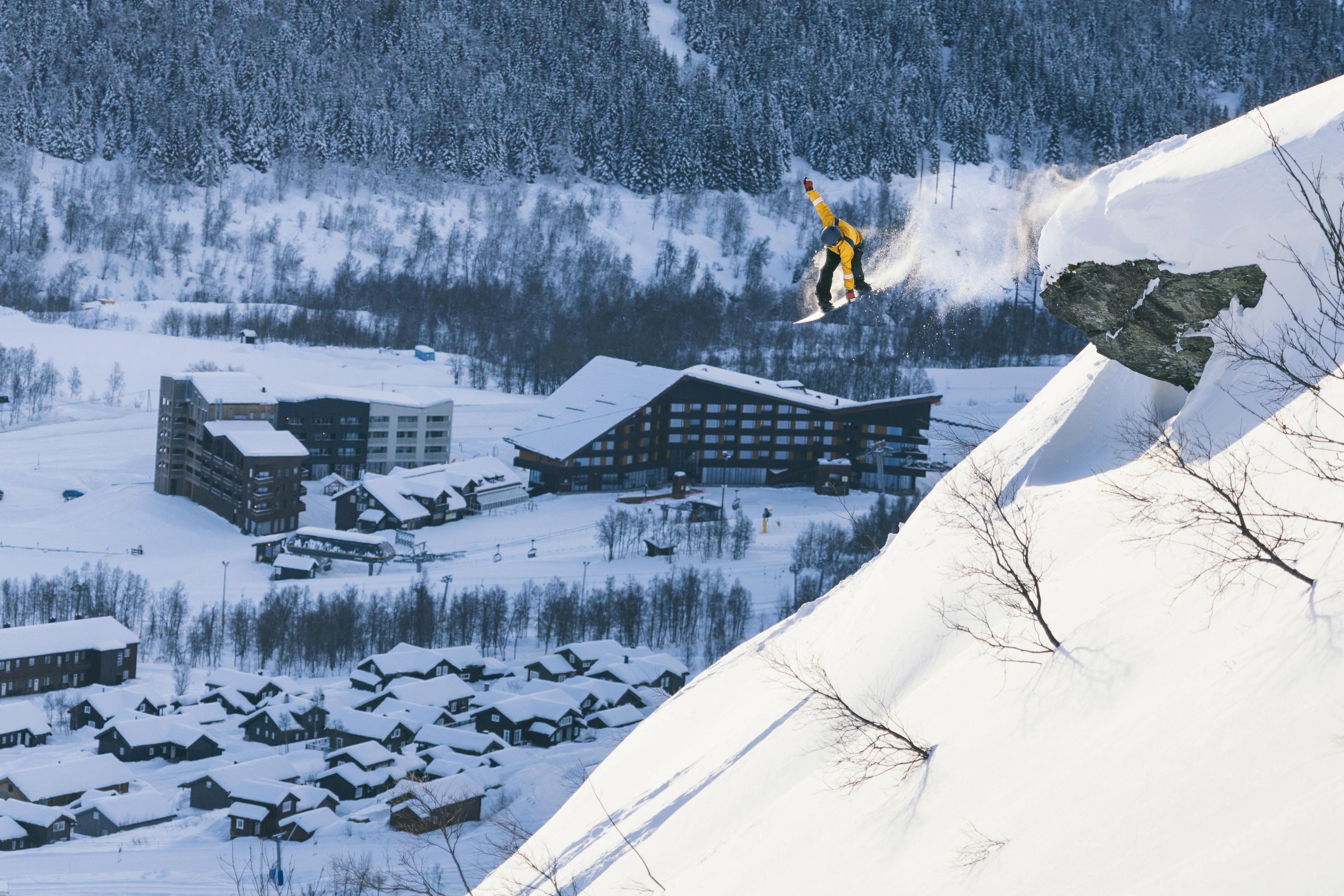 A person jumps off a cliff on a snowboard with Myrkdalen Hotel in the background.