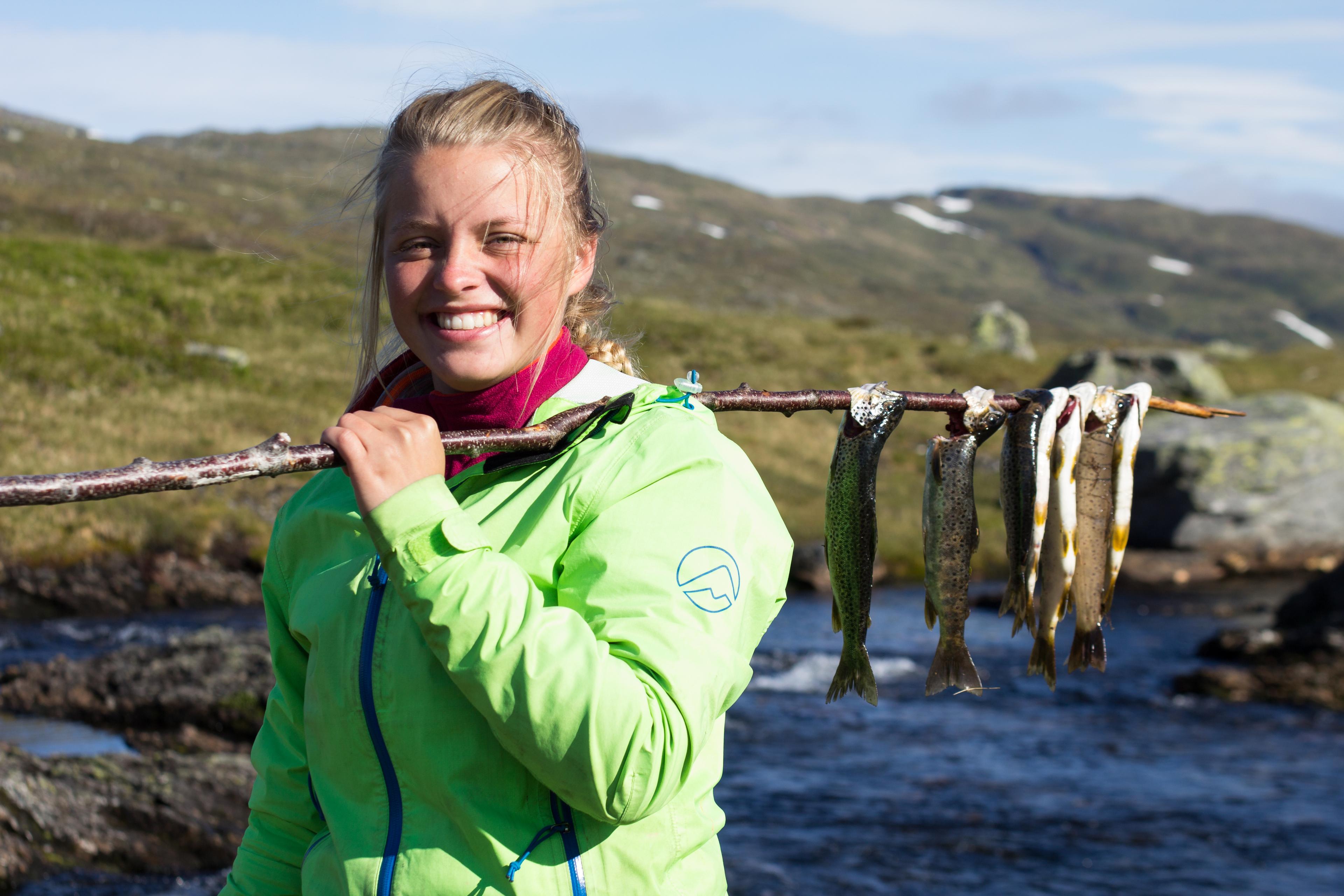 Smilende jente ved et fjellvann viser frem fiskefangsten på en stokk som hun bærer over skulderen.