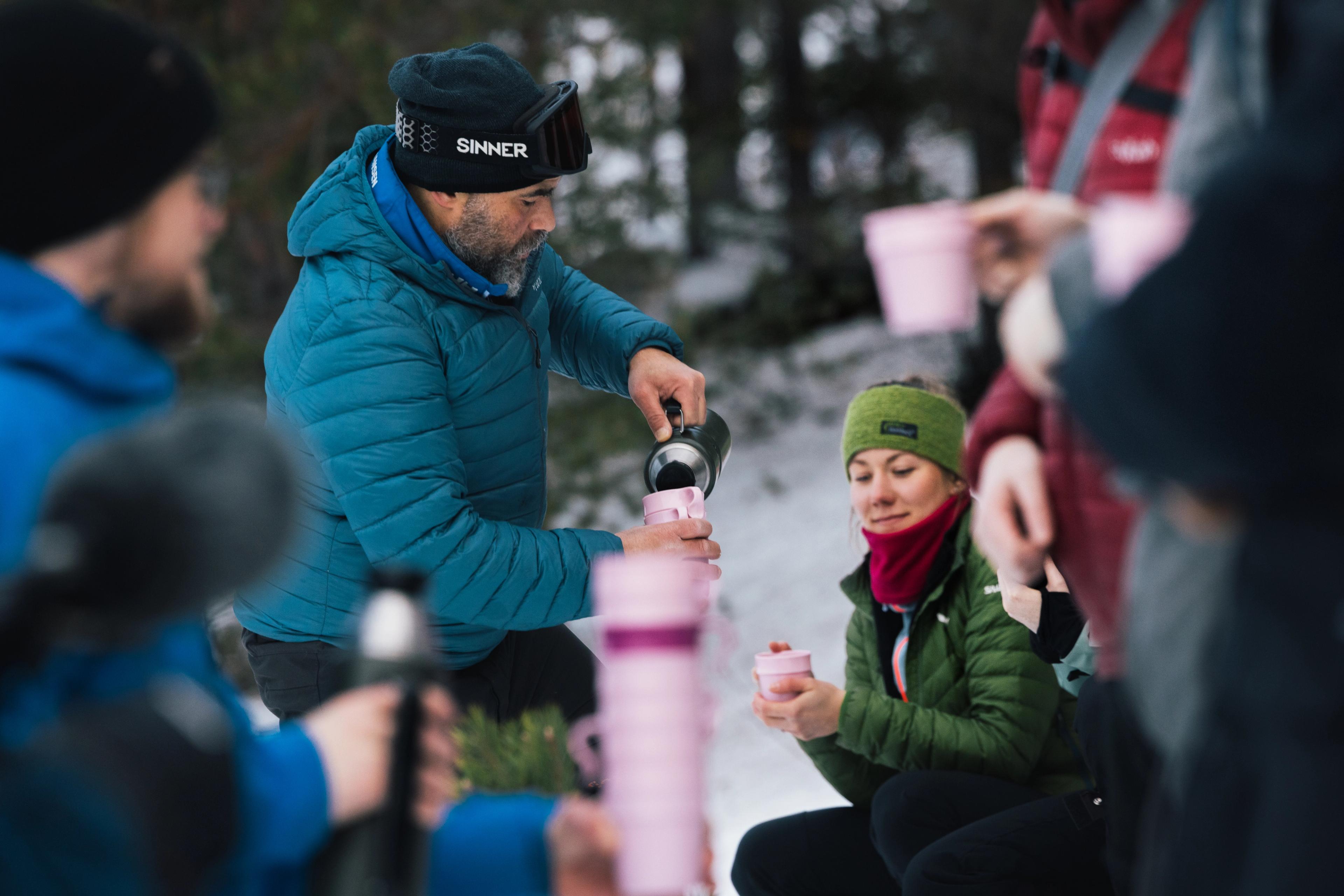 A man in a blue down jacket serves coffee from a thermos to winter-clad people on a snowshoe trip.