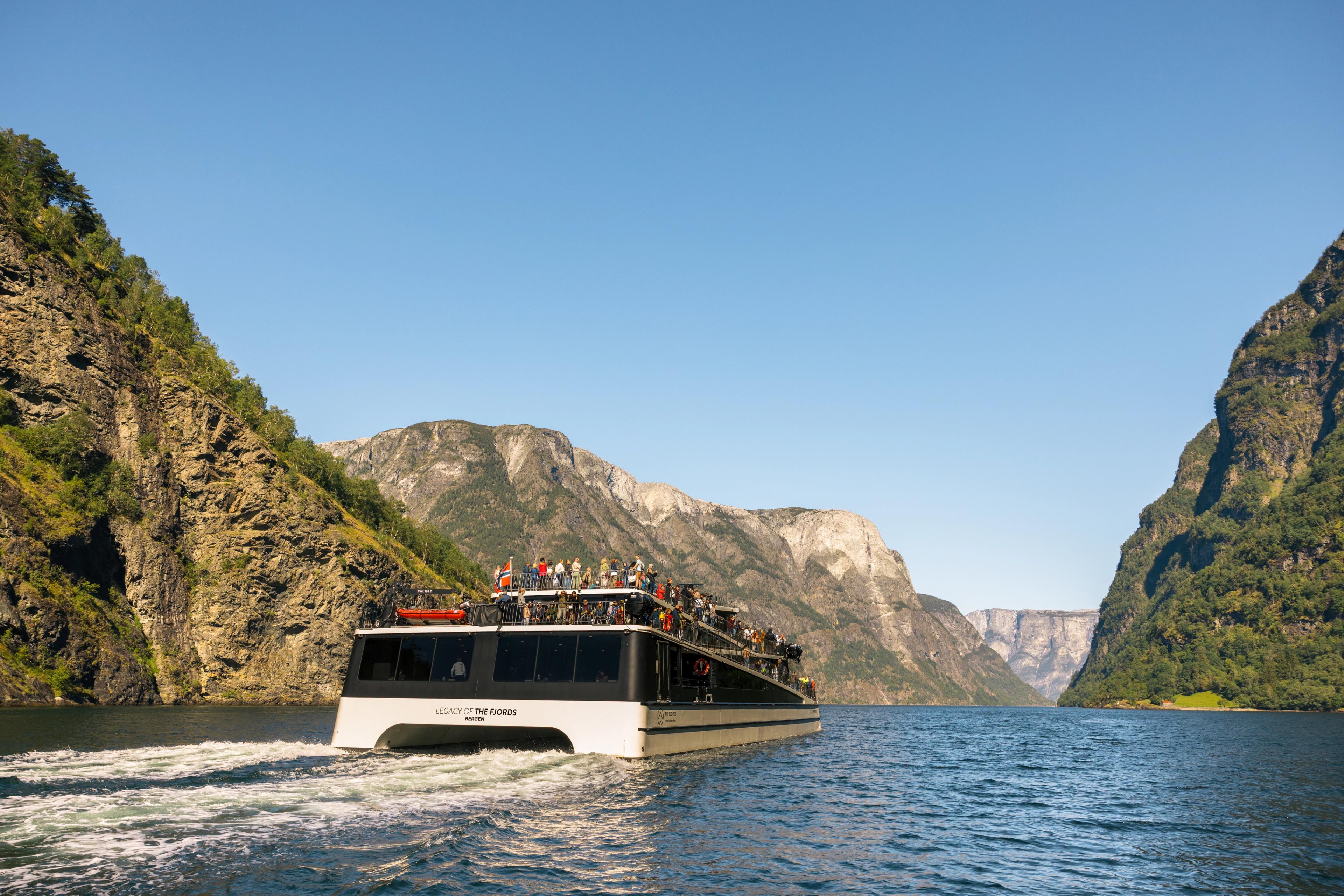 Katamaran segler inn i Nærøyfjorden med de bratte fjellsidene rundt. Tak og dekk er fyllt av folk.