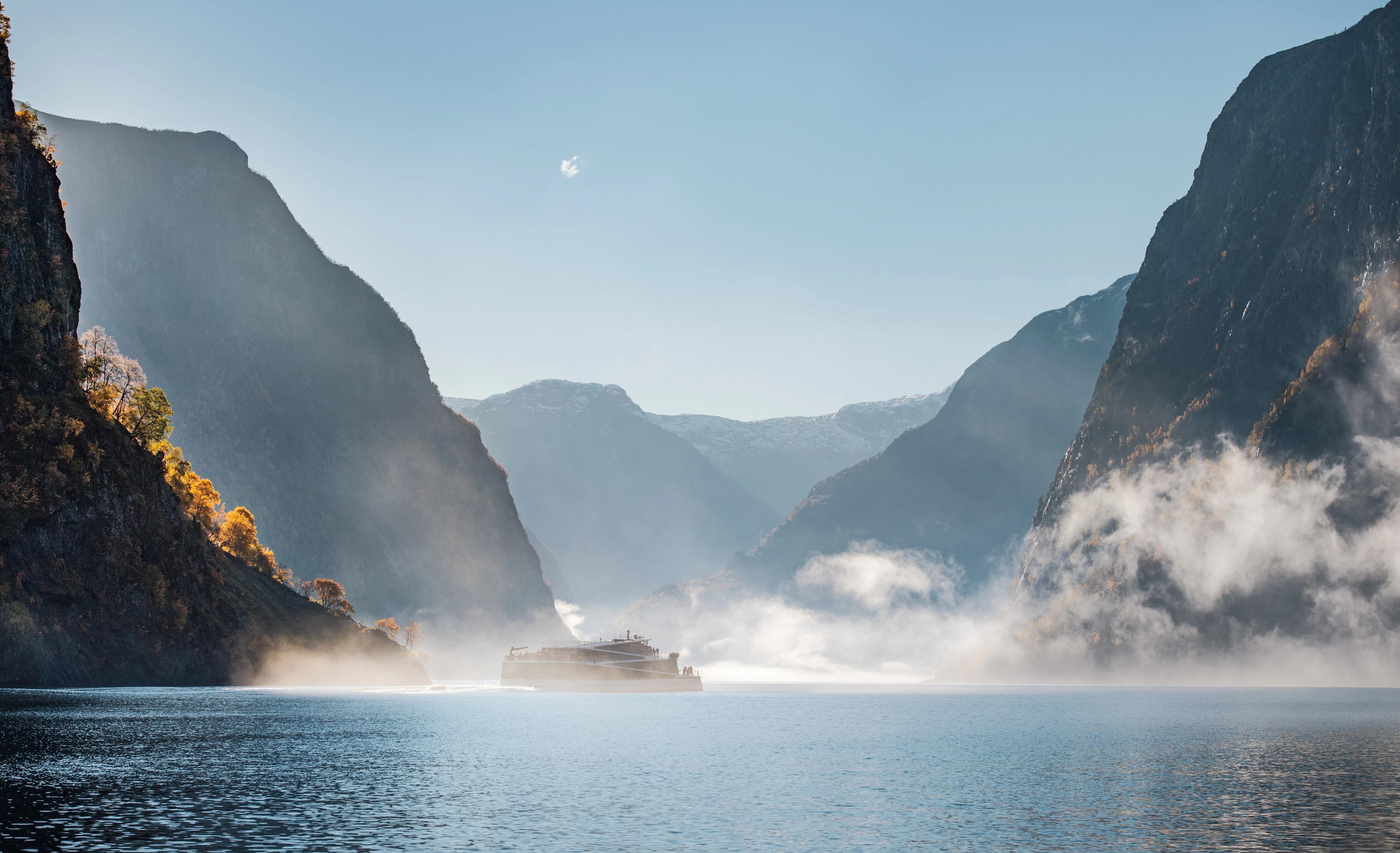 En båt omgitt av lett morgentåke som seiler i Nærøyfjorden om høsten. Solen skinner på oransje trær i fjellsidene.