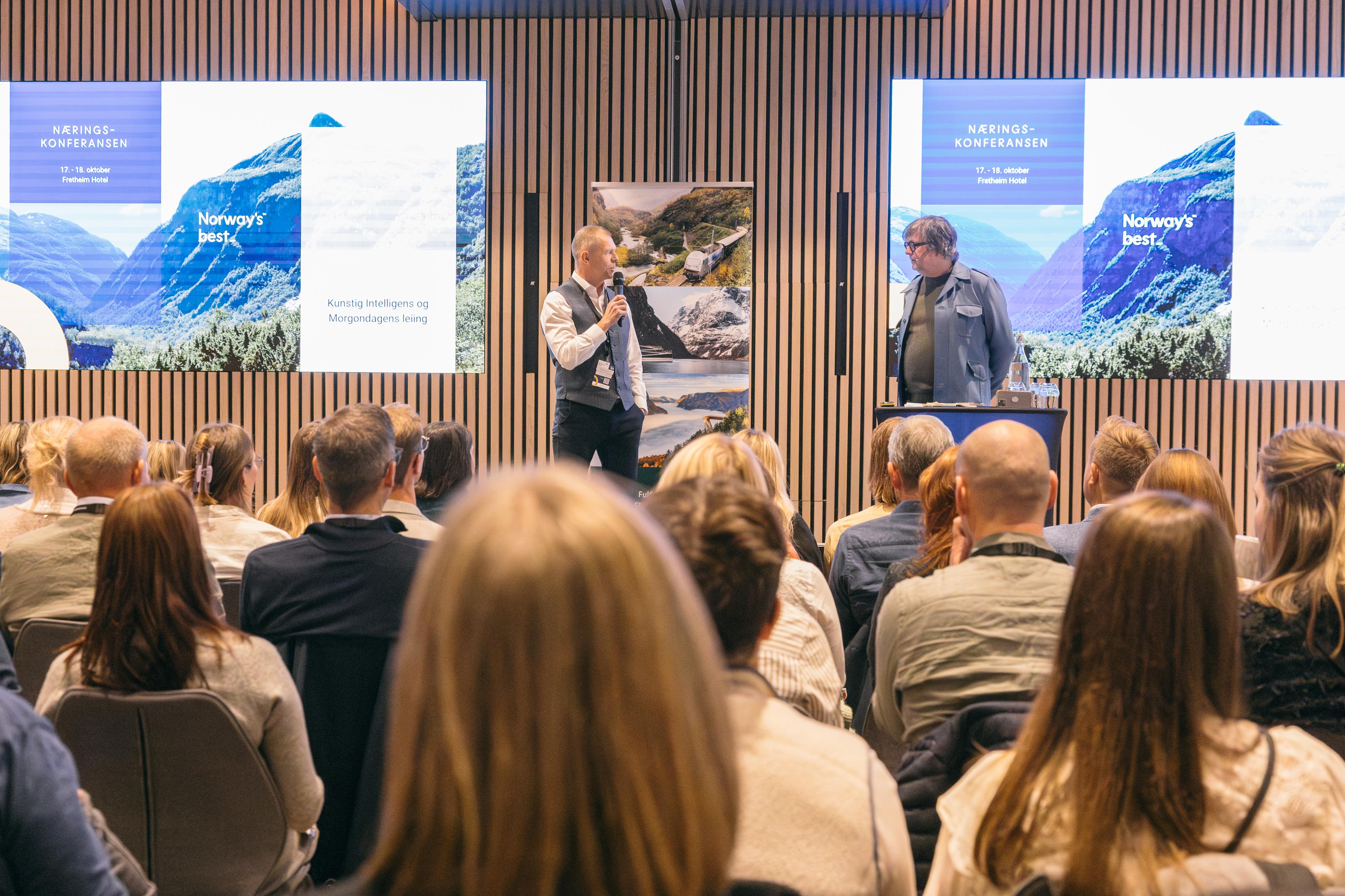 Foredragshaldar i samtale på scena under Næringskonferansen, med publikum i salen og presentasjon på skjerm bak.