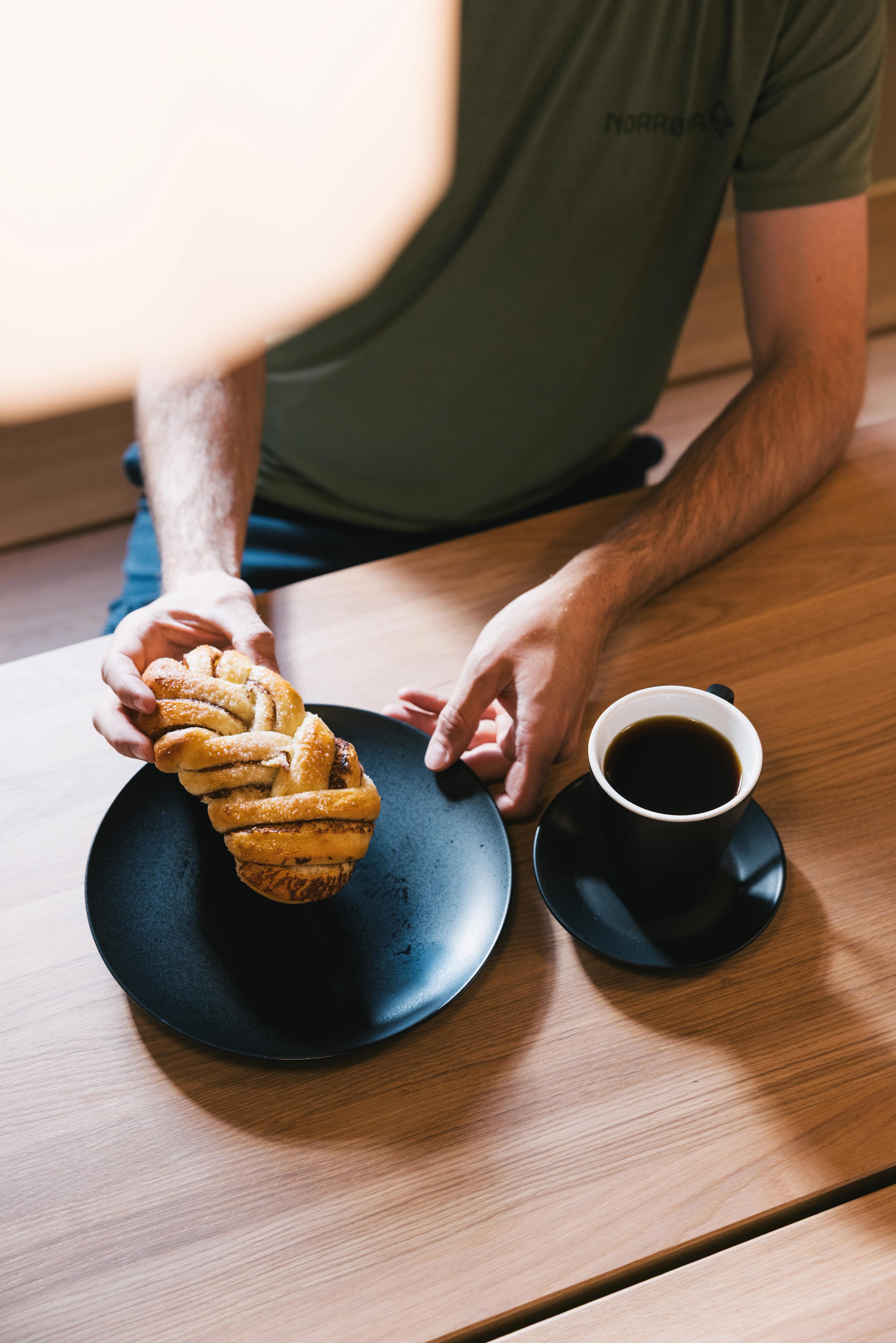 Die Hände eines Mannes heben eine Zimtschnecke hoch. Untertasse und Kaffeetasse auf dem Tisch.