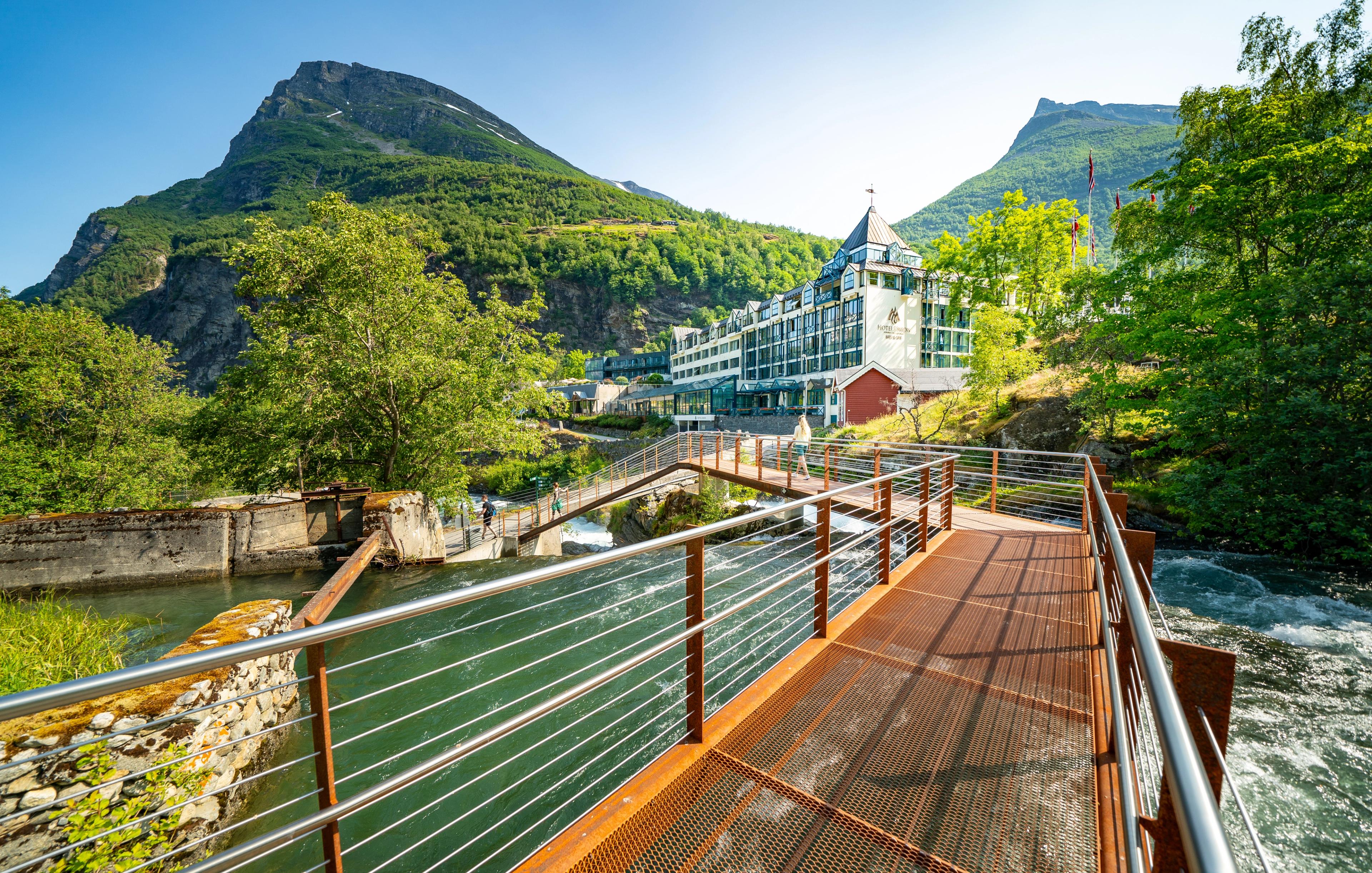 A pedestrian bridge crosses a river, with a hotel and tall mountains in the background.