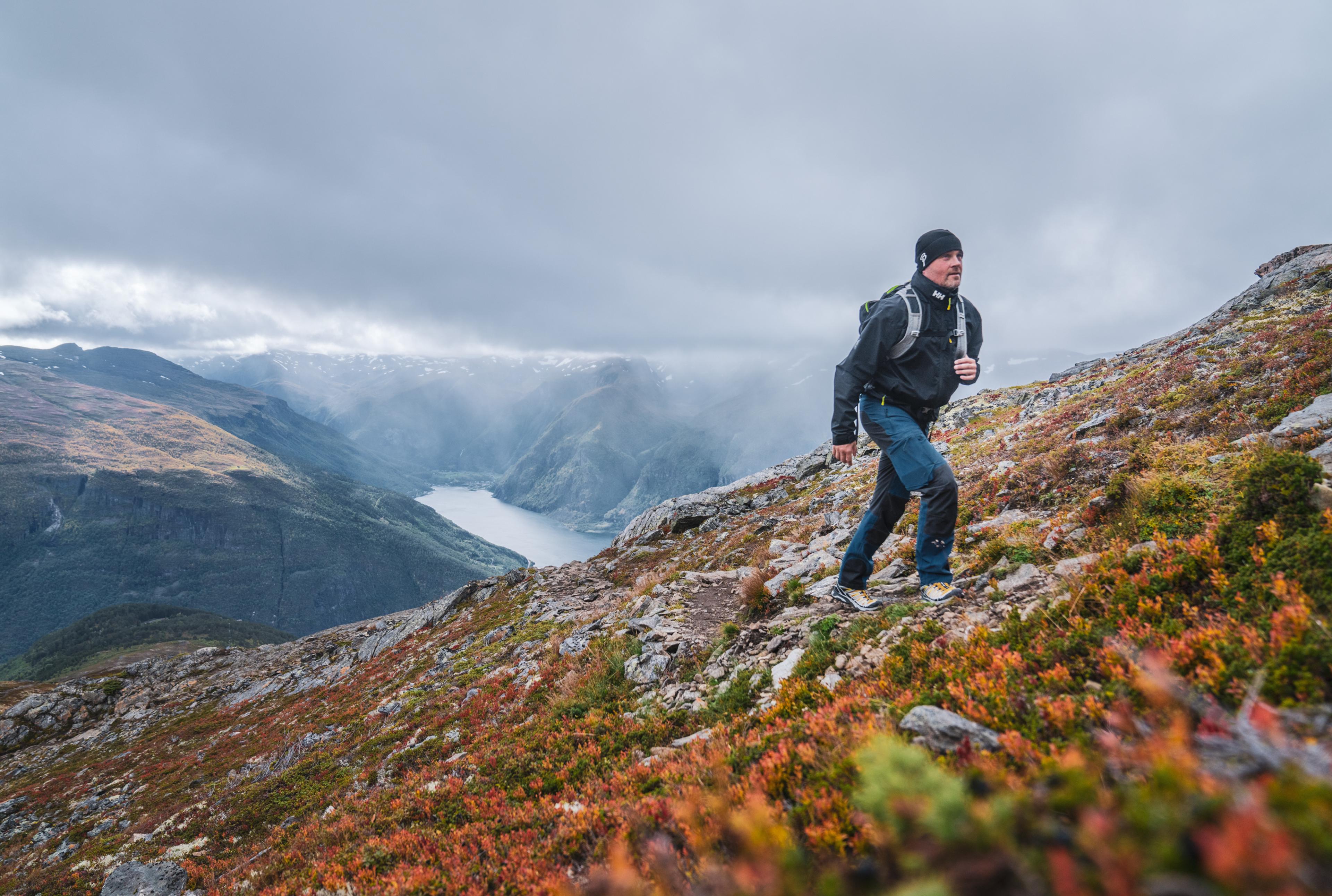 Ein Mann, der einen Hügel hinaufgeht mit der Fjordlandschaft, die dahinter im Nebel liegt.