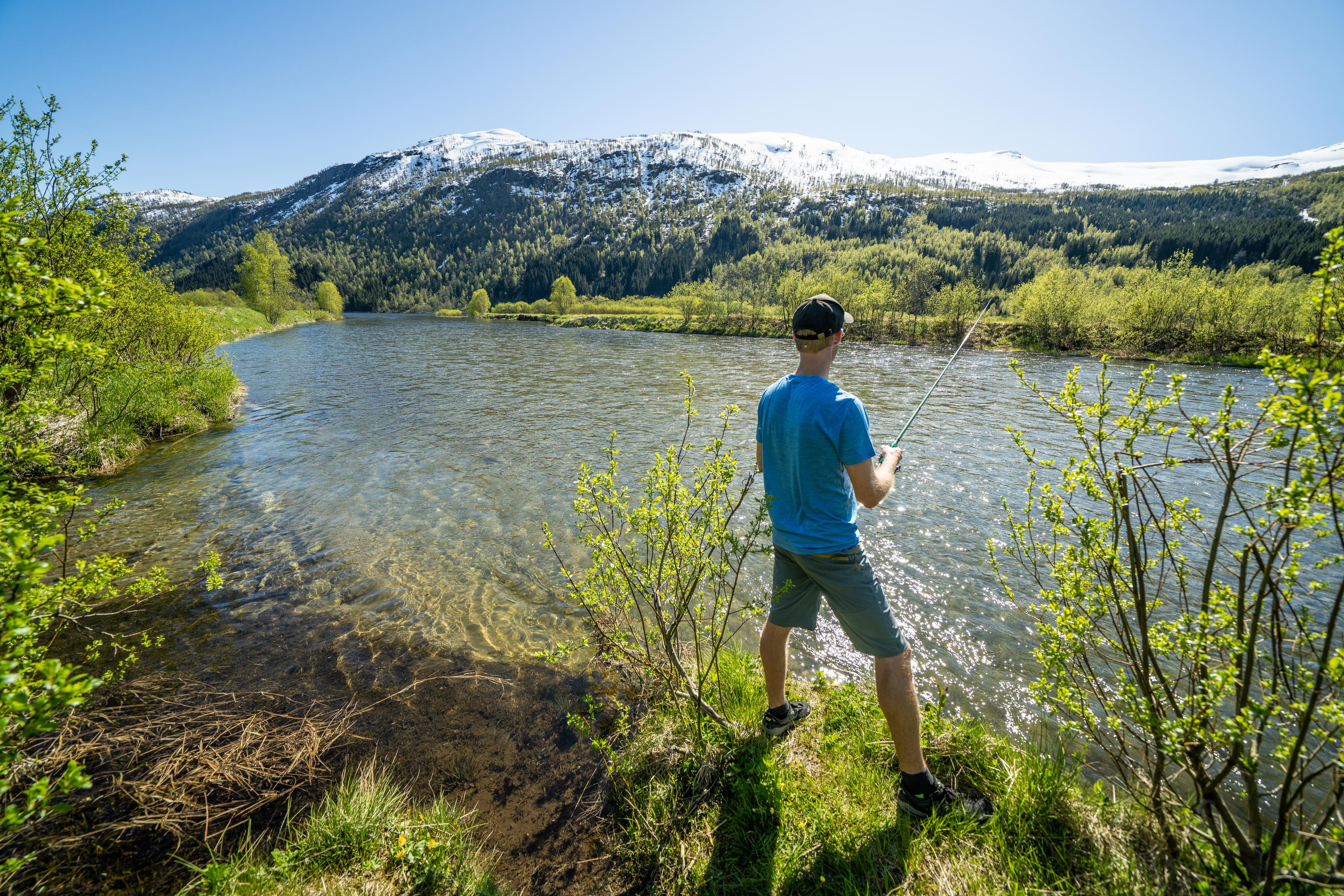 Mann fisker i et fjellvann en sommerdag. Det er fremdeles snø på fjelltoppene.