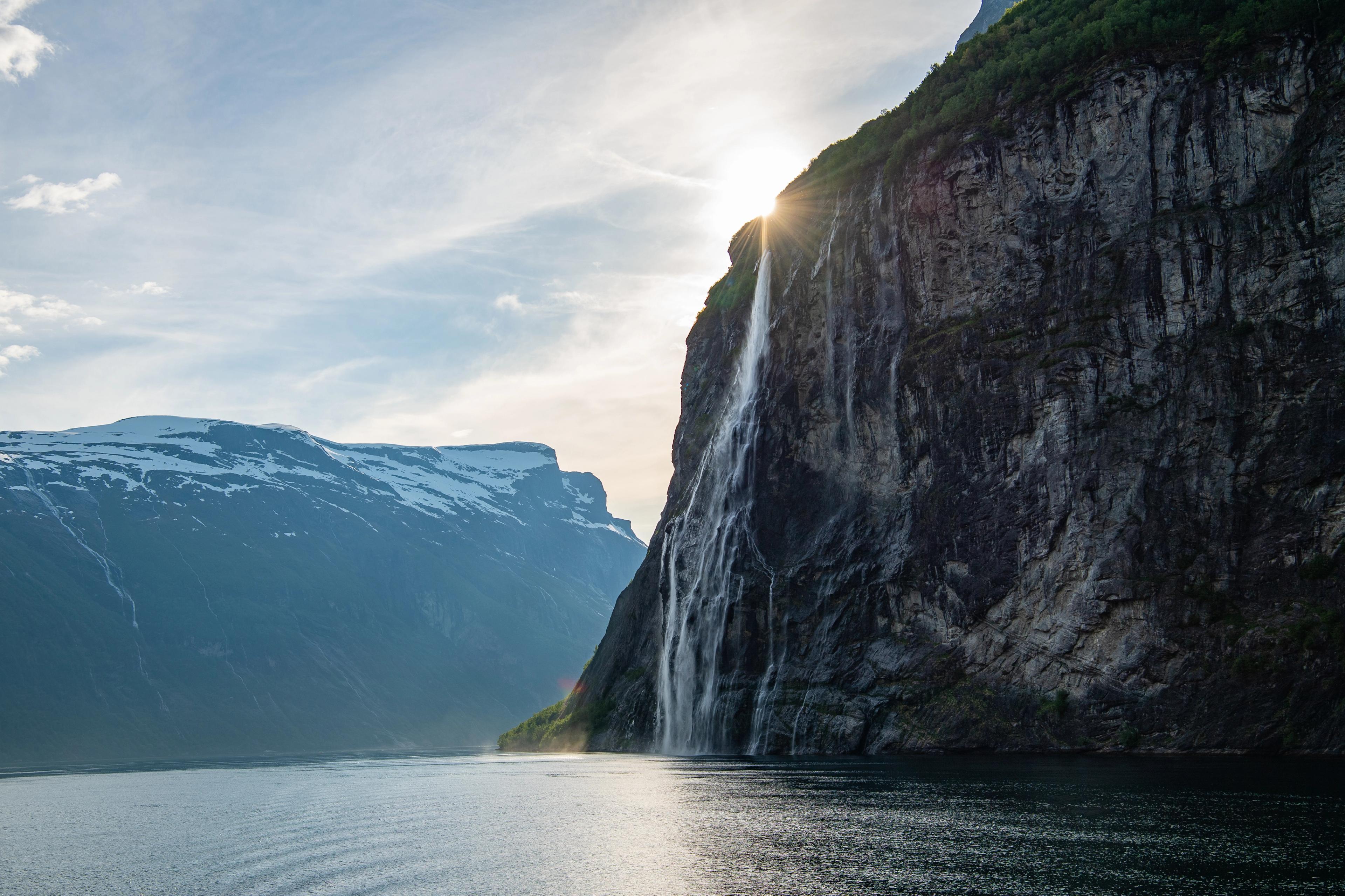 View of Geirangerfjord with a steep mountainside and a waterfall plunging into the fjord. The sun peeks over the mountain.