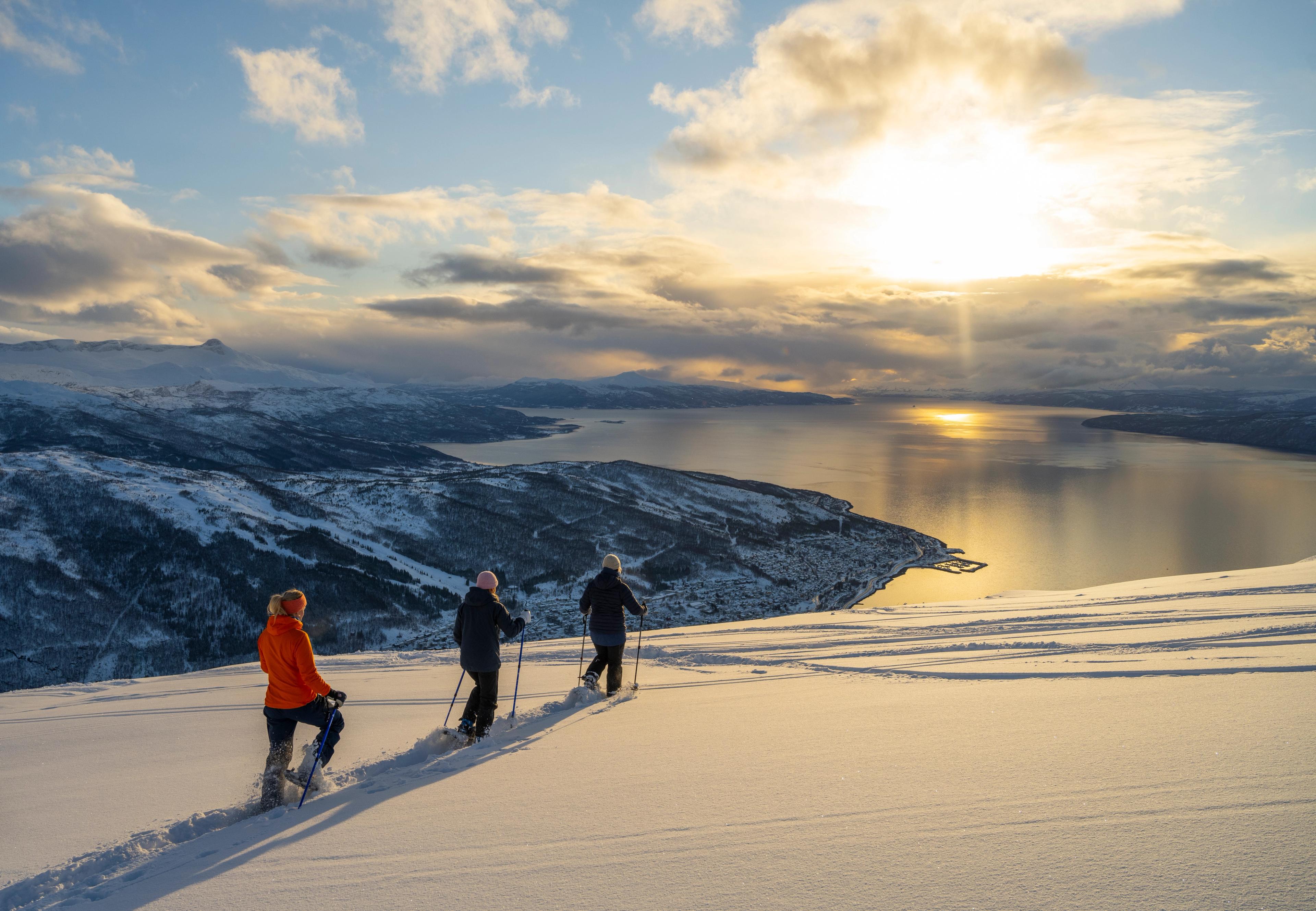 Three people snowshoeing down a mountainside with views of the fjord and the low winter sun.