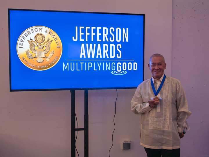 Mike Feria aka Dr. Mauro Feria Tumbocon, Jr. on the photo standing beside a flat screen tv with texts JEFFERSON AWARDS Multiplying good