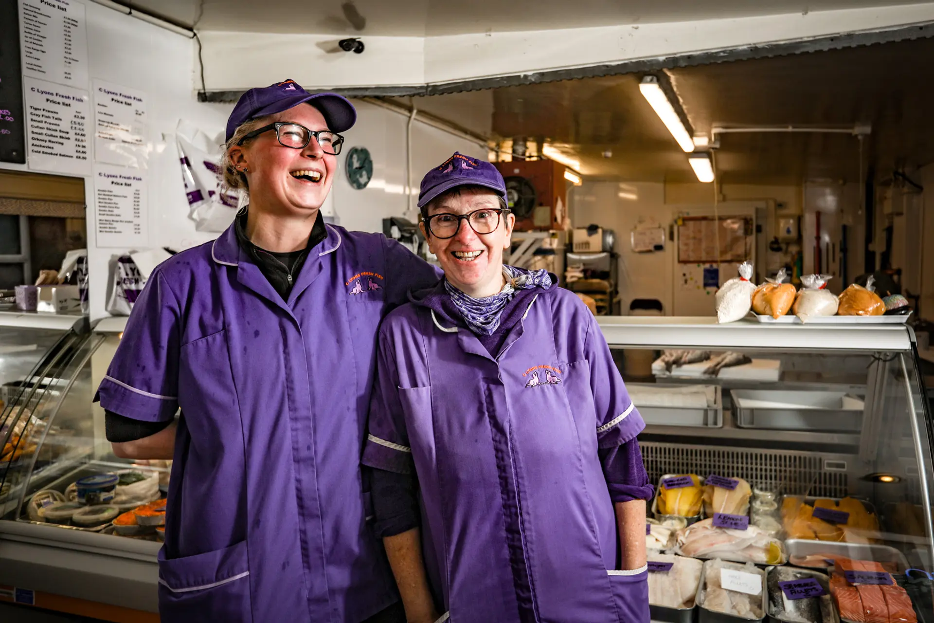 Two women wearing purple uniforms are standing close to each other and laughing at the camera, they are inside a fish shop. 