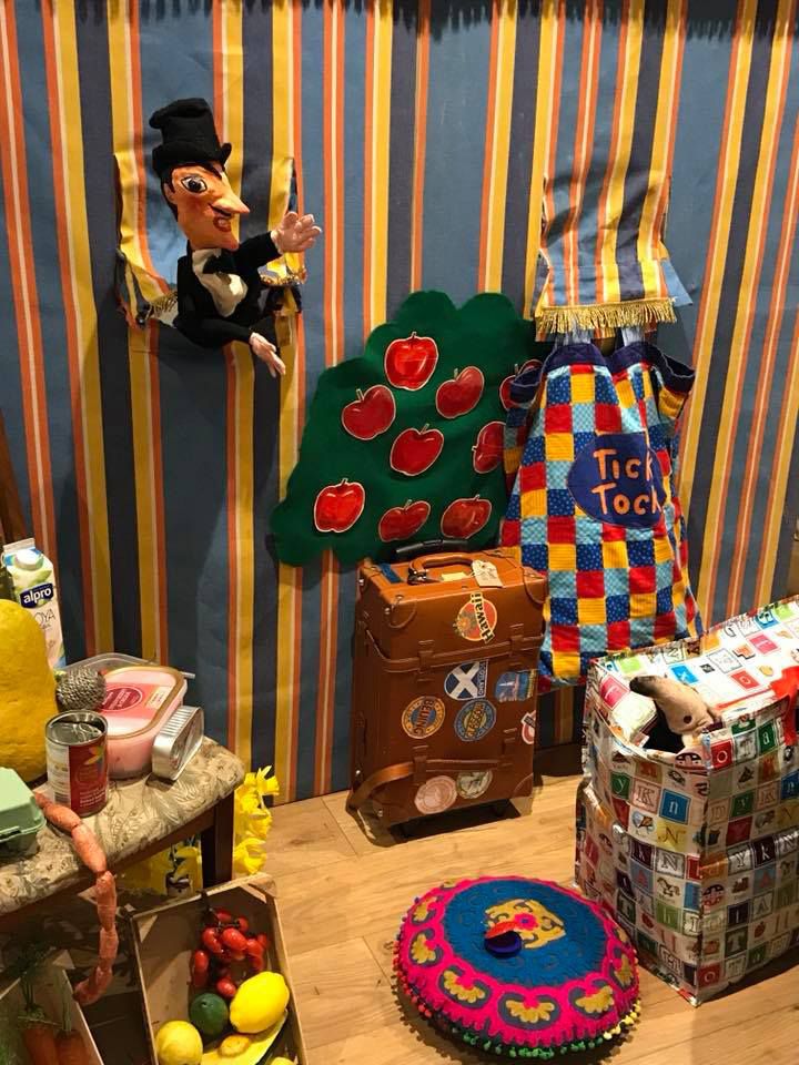A busy, colourful picture. There’s a section of a striped puppet booth, with a moustached papier-mâché puppet wearing a top hat and bow tie, popping out. Lots of props and objects beginning with the sound ‘a’ are laid out, for example an aardvark and an apple tree.