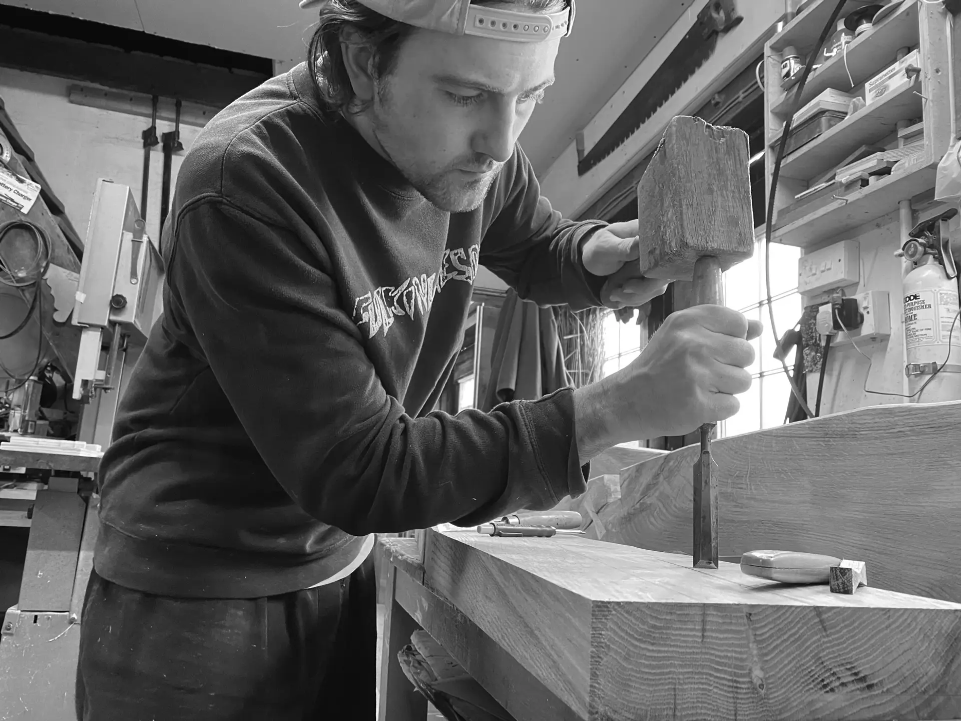 Man in his twenties, with a baseball cap on back to front, holds a wooden mallet and tool and works on a smooth, thick plank of oak wood. He is inside a workshop.