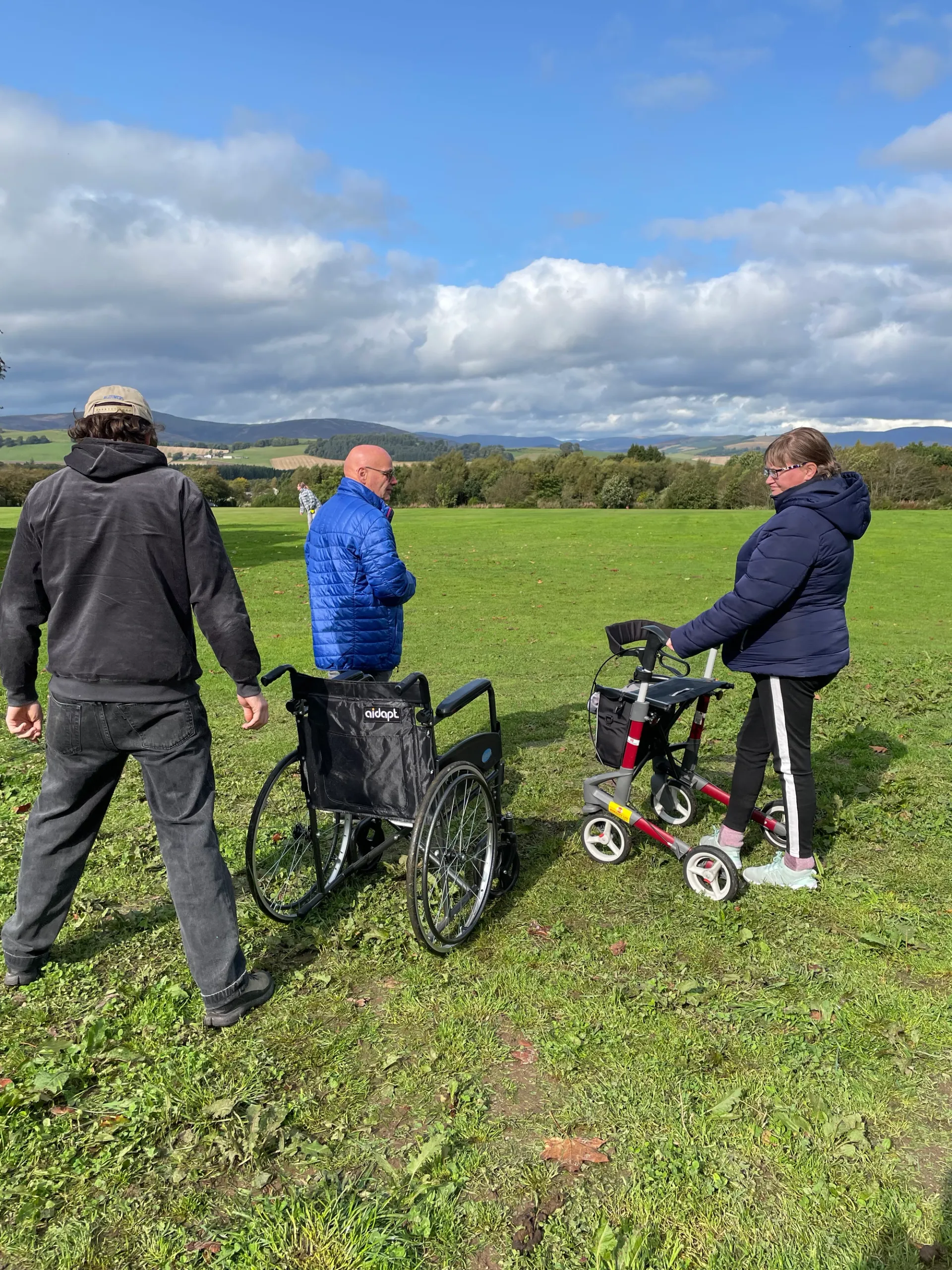 3 people stand looking at a huge sweep of green grass, trees and hills under a blue, white and grey cloud-filled sky. One person stands with a mobility walker, 2 people stand next to an empty wheelchair.