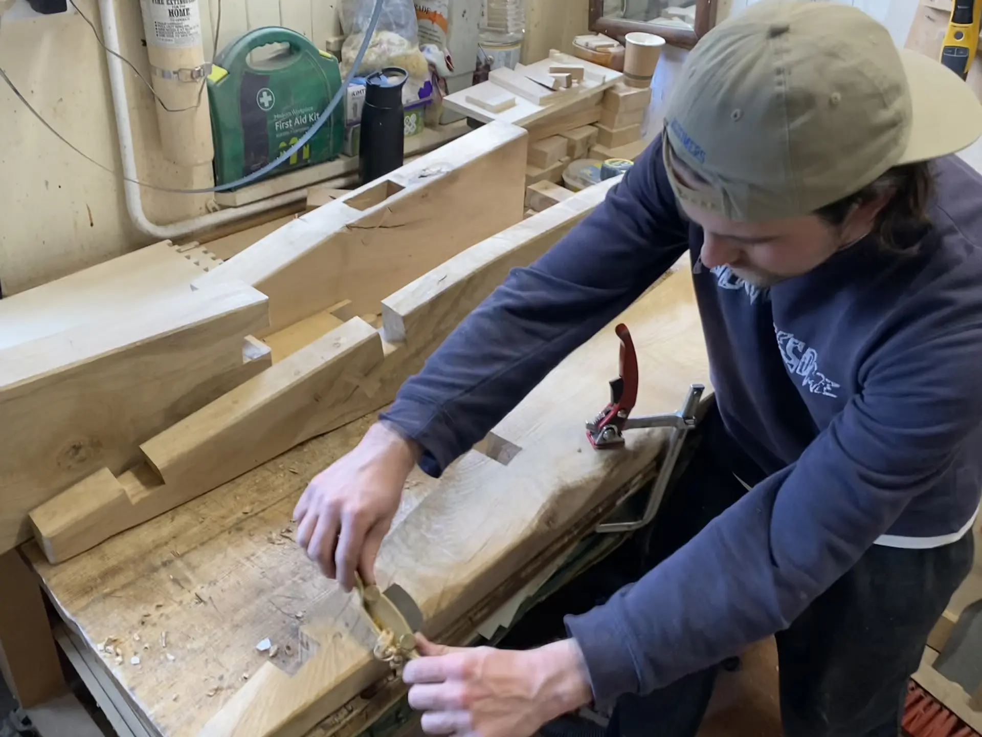 A man works with large pieces of wood inside a workshop. He wears a baseball cap back to front. He is using both of his hands outstretched, to work the wood with a tool. There are pieces of wood cut into various shapes and with sections cut out of them, large slots