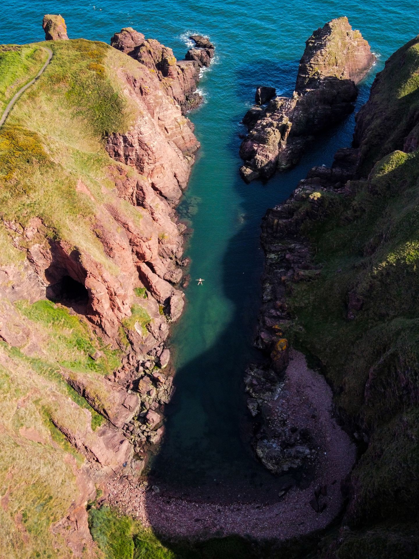 A drone shot looking directly down at a coastal cliff scene. There is a large U shaped inlet filled with blue water which is surrounded by vibrant green grass and maroon coloured cliffs. In the centre of the sea is a small person floating on their back like a starfish.