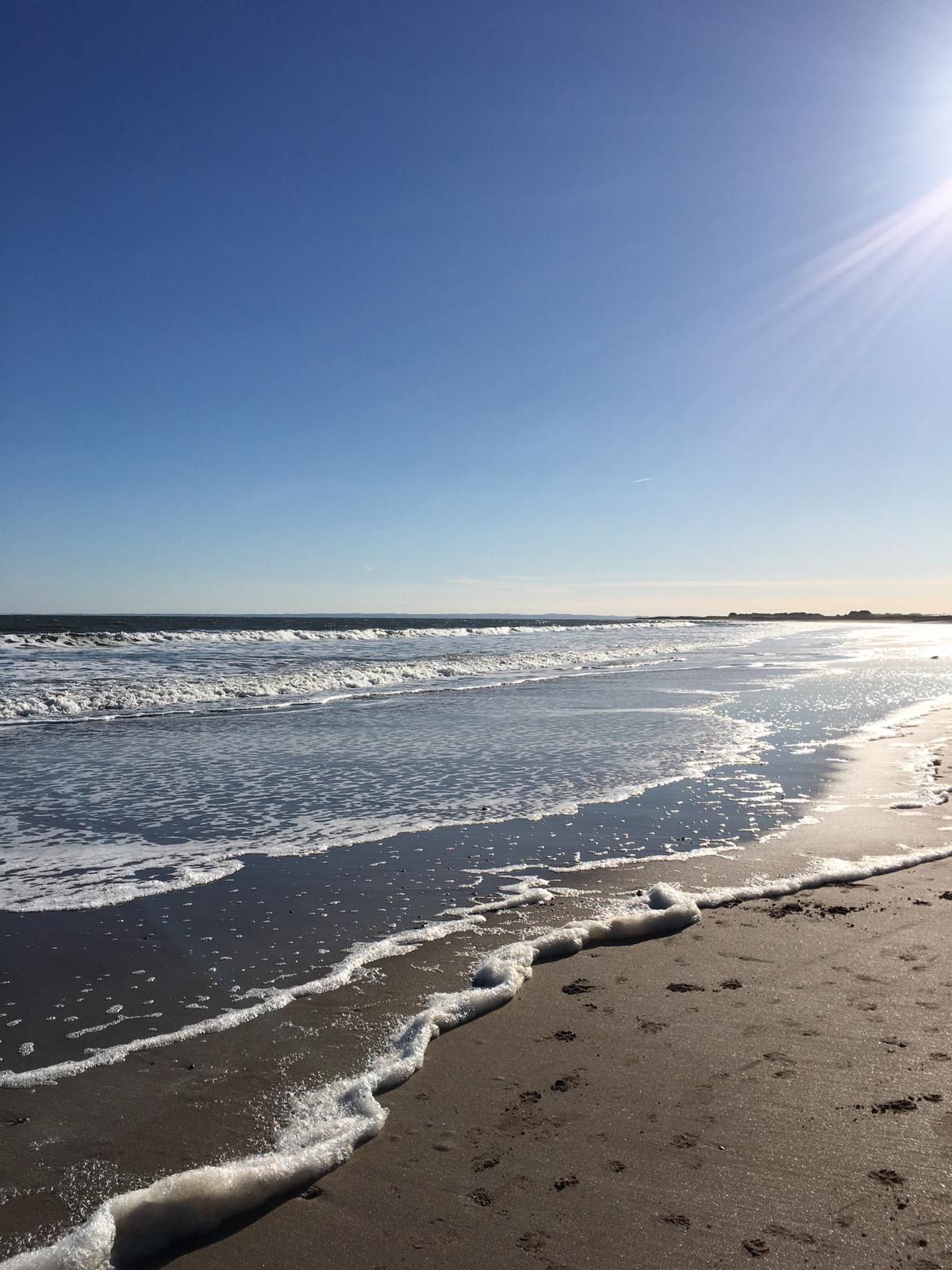A photo of Arbroath beach looking towards the horizon to the right. Taken late afternoon on a spring day with clear blue skies and frothy waves, with footsteps on the sand in the foreground.