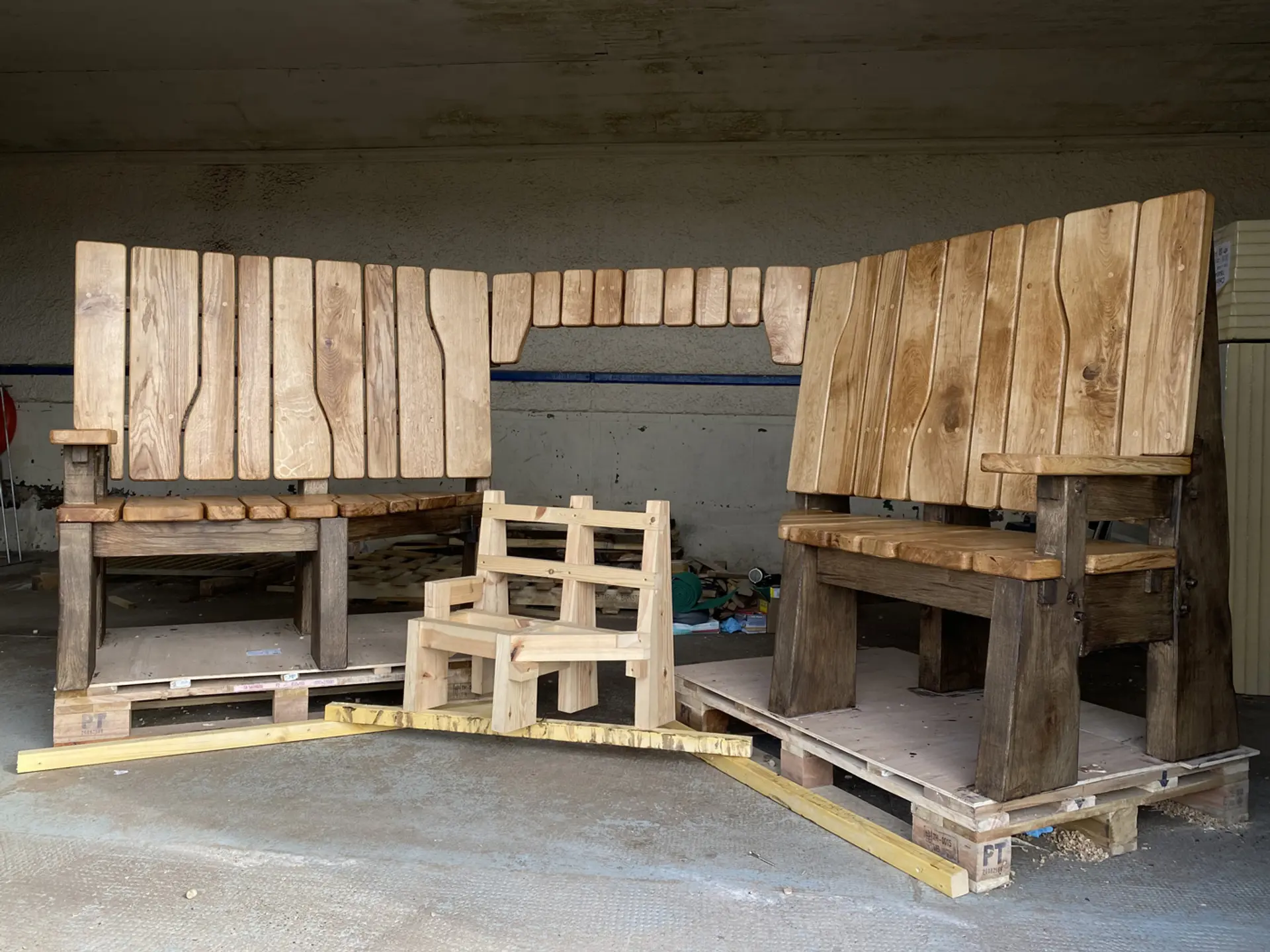 Inside an empty-looking shed or storage place a large, fully built oak bench sits behind a much smaller version of the same bench. They are raised up on large wooden palettes.