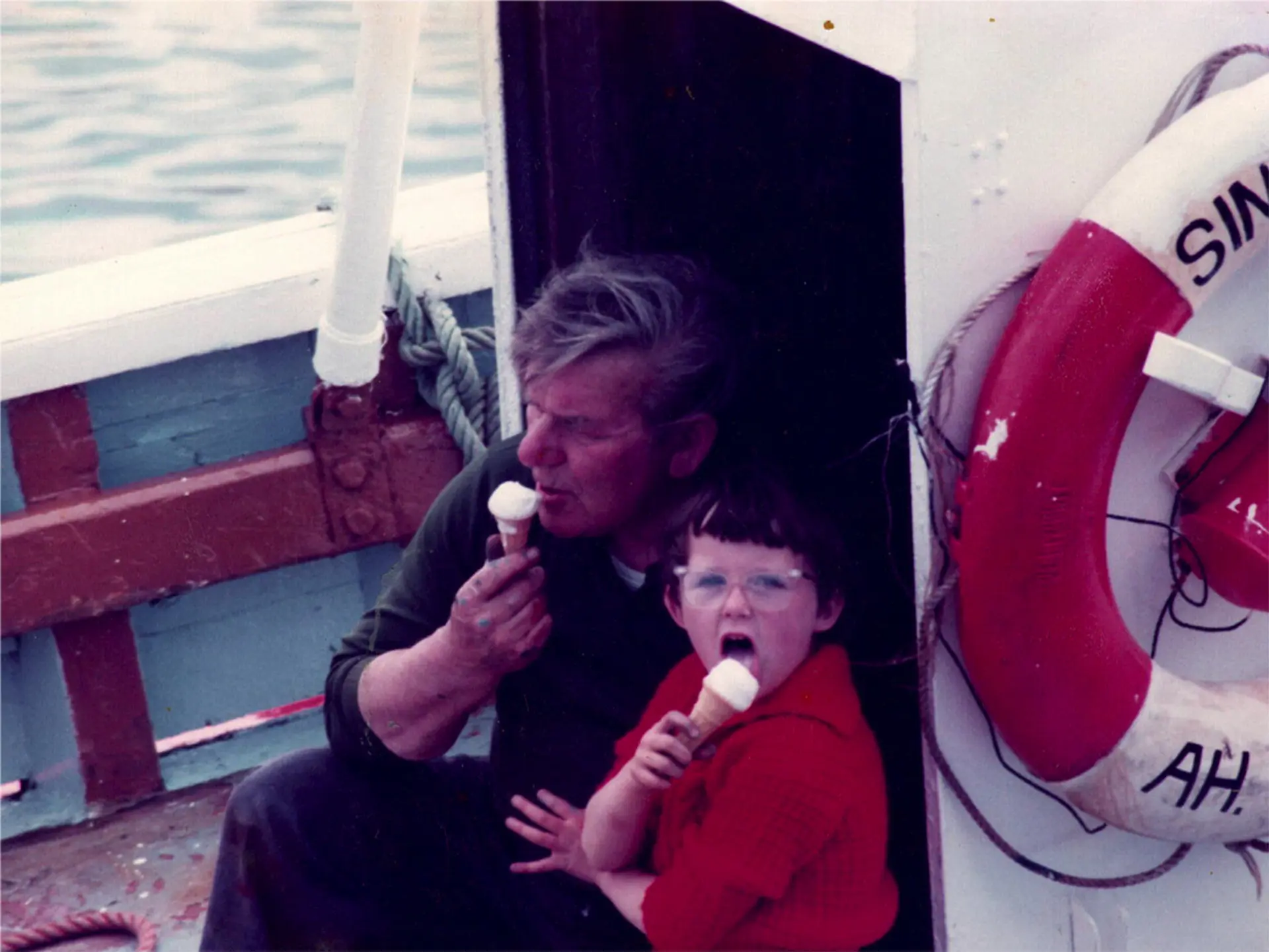 An image from the past, a man and child are eating ice creams in cones on a fishing boat. We can see a red and white lifebuoy.