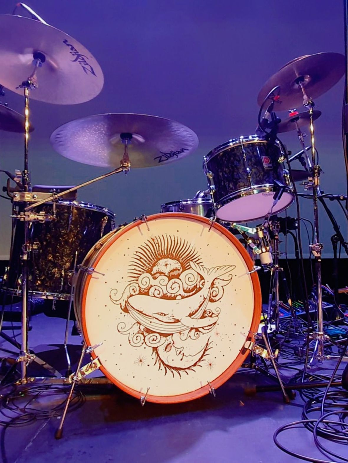 A black drum kit on a stage in a theatre with a whale mandala image on the front of the bass drum.
