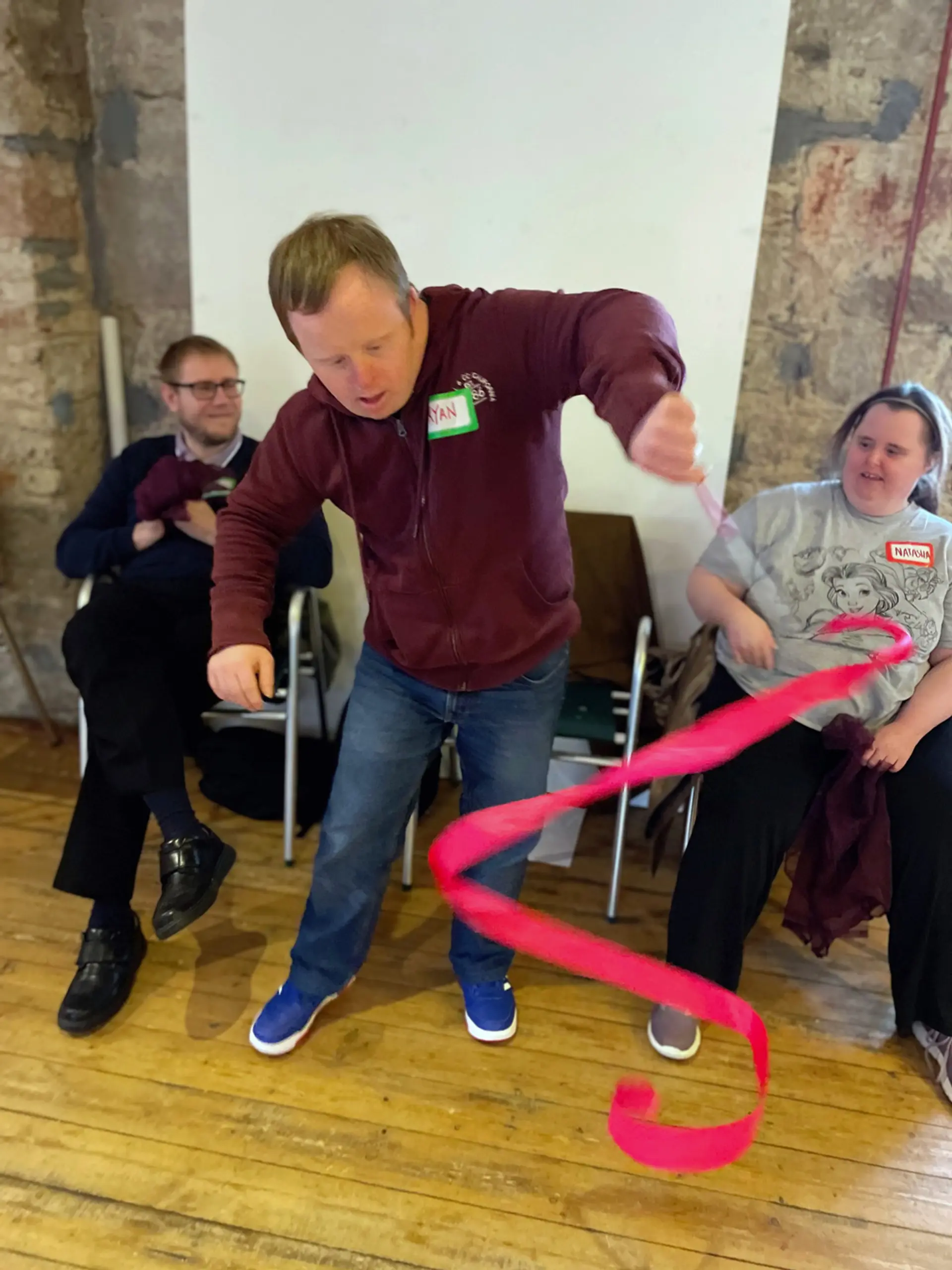 Three people are at a drama workshop. The person in the centre has a name tag on which says ‘Ryan’. He is standing up and holding a bright pink ribbon on the end of a stick, he is spinning it to create a spiral shape. The other two people in the picture are seated and look entertained.