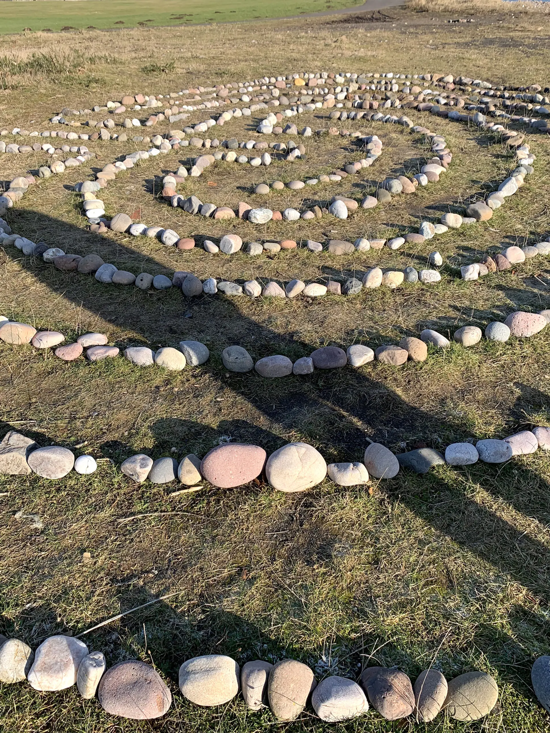 -Arbroath COVID-19 stone spiral A collection of stones arranged on grass in a circular pattern. It is sunny and a person casts a shadow across the image. 