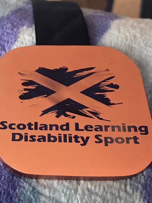 1 bronze medal with a dark blue ribbon and 1 white medal, also with a dark blue ribbon. They both have a Scottish flag pattern and the words ‘Scottish Learning Disability Sport’ on them.