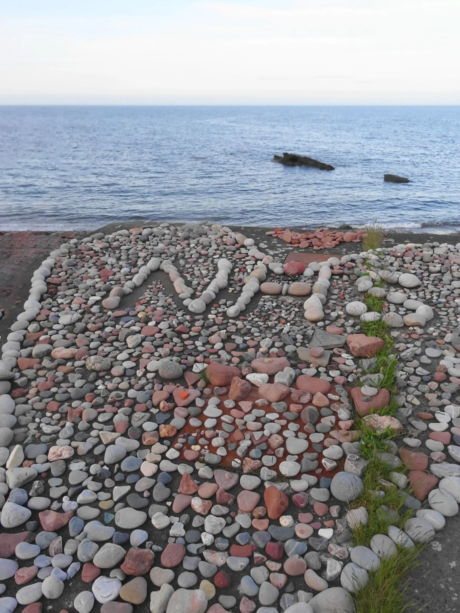 A large collection of beach stones arranged in a love heart with ‘NHS’ written in the centre. In the near distance we can see the blue sea. 