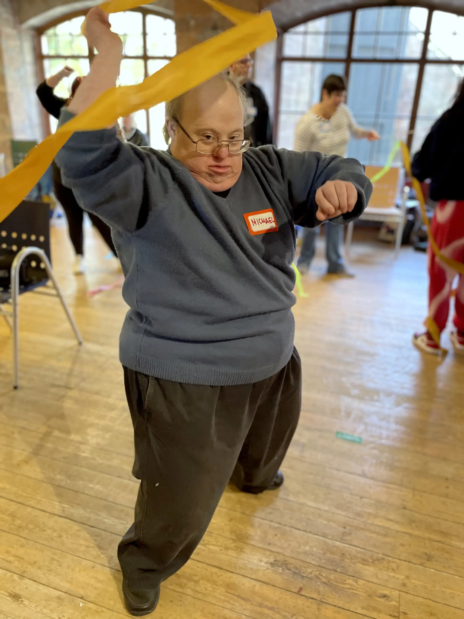 A person with a name tag which says ‘Michael’ on it is at a drama workshop. He wears glasses and has his arm above his head, he is spinning a ribbon attached to the end of a stick to create a spiral shape.