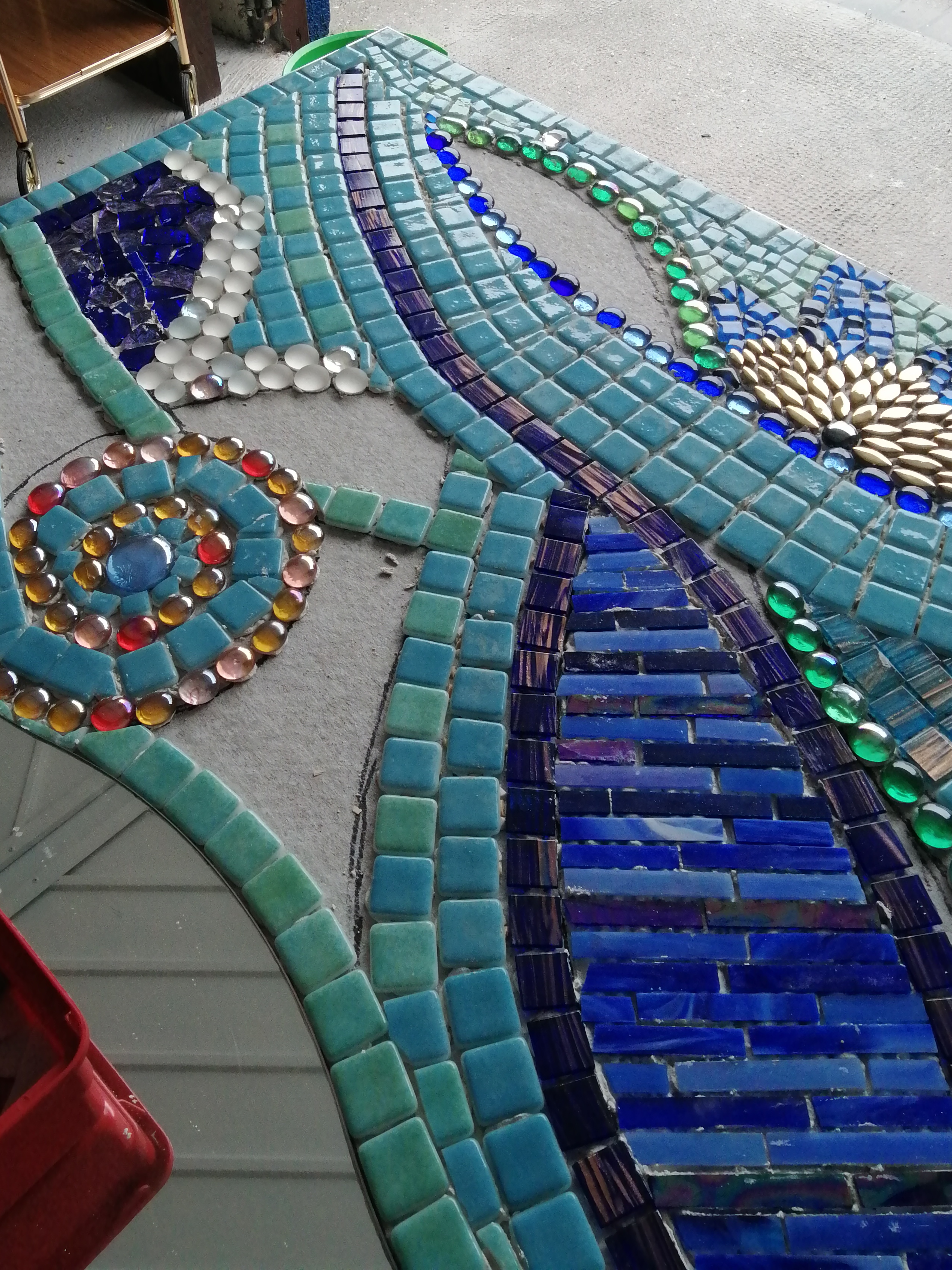 An abstract patterned mosaic lockdown project, made with little green and blue tiles interspersed with swirls of glass beads in various colours, in the background is a section of a garage with some bit of furniture and the tiled ground.