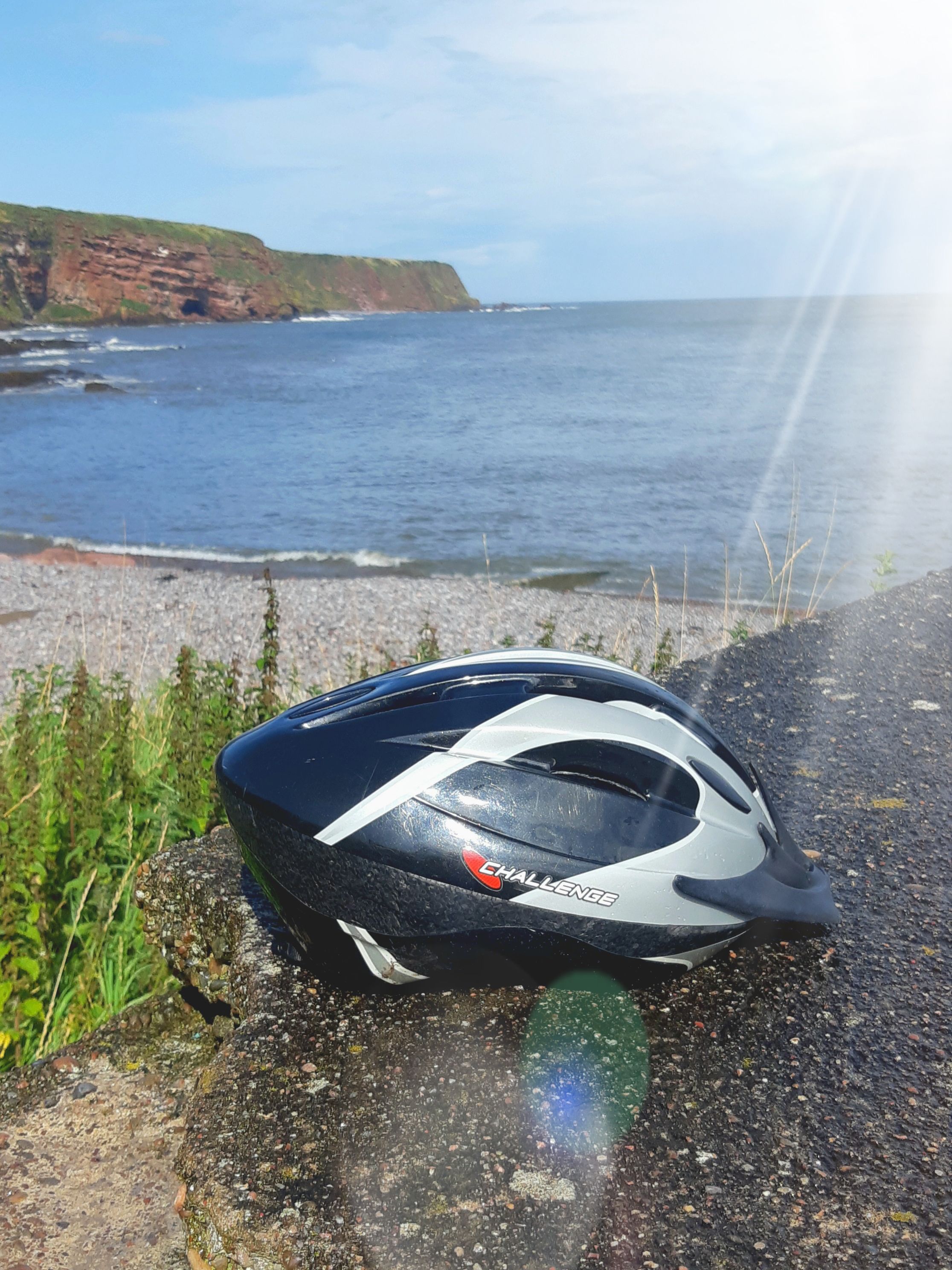The sun shining down from a blue sky onto a beautiful coast line and a black slimline bike helmet sitting on a rock.