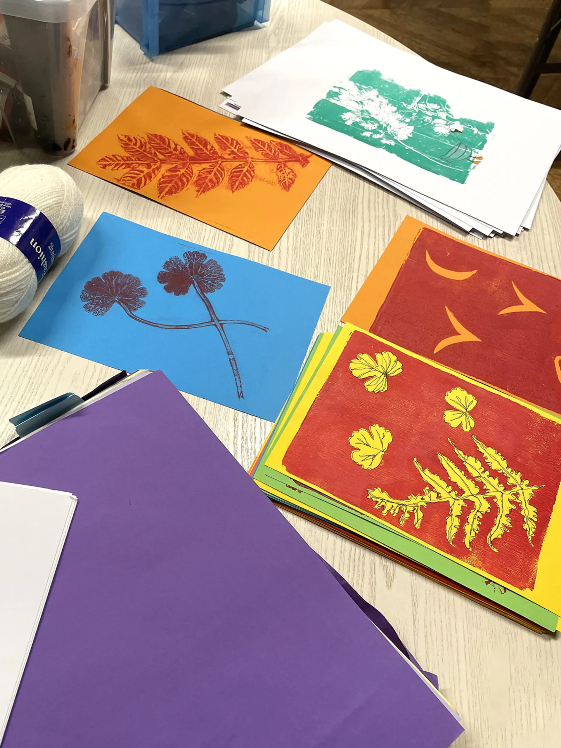 5 pieces of artwork created by printing leaves with ink and displayed on coloured paper on a table top.