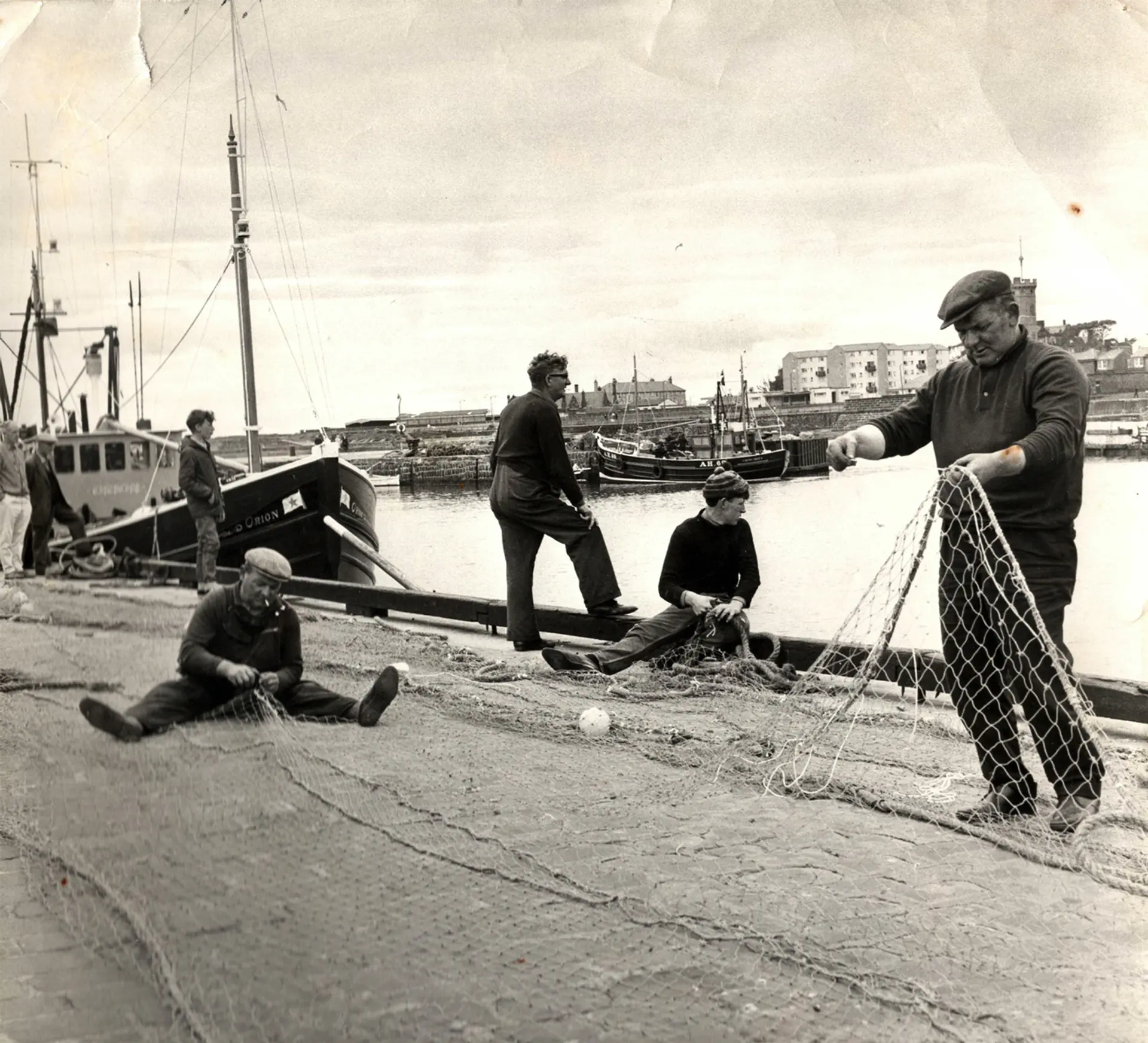 A sepia image from the past. Seven fishermen are standing and organising their nets in a harbour. They are of varying ages.